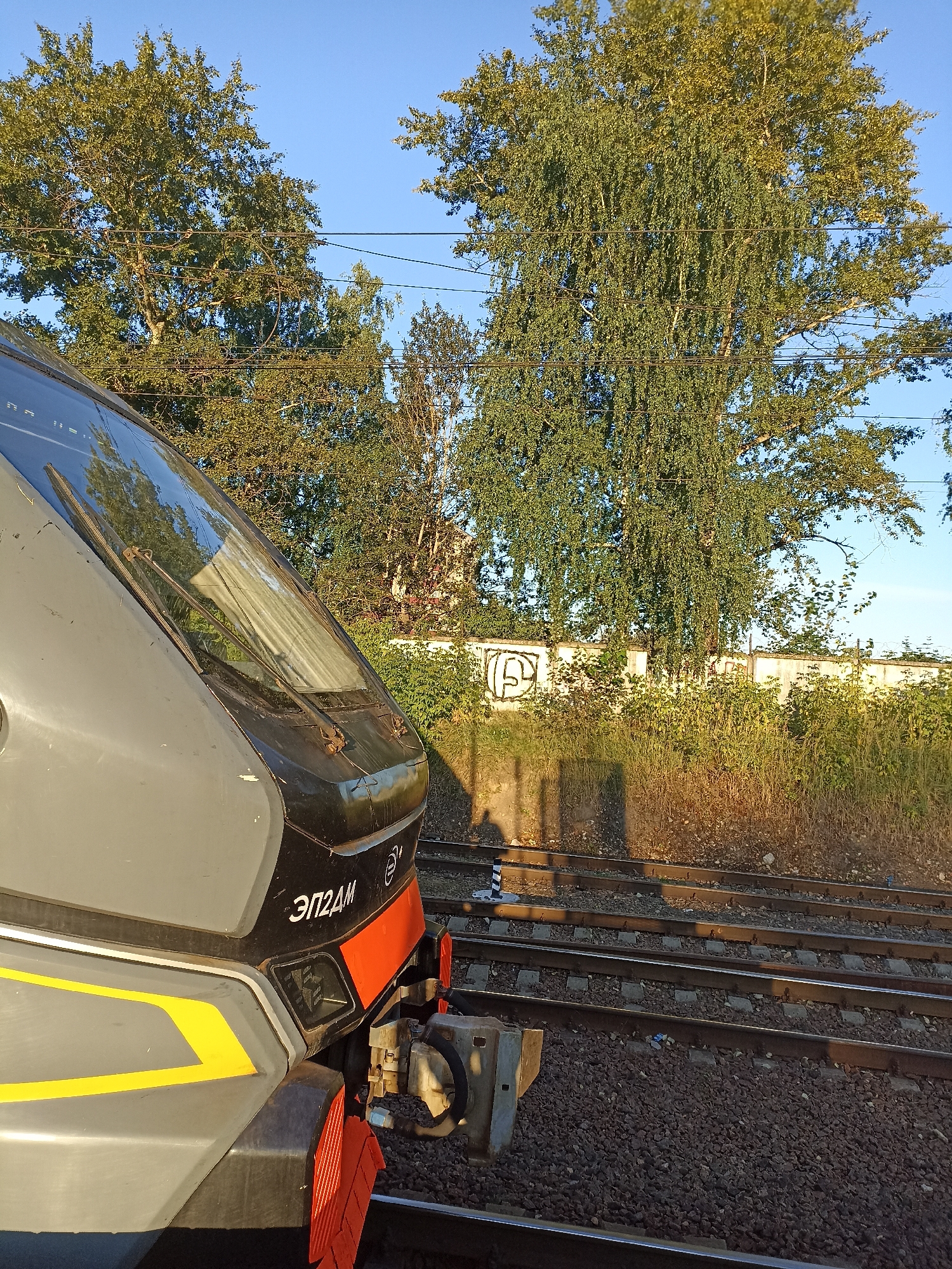 In Chekhov, Russia, a close-up shot focuses on the front of a modern gray and black train, identified by Cyrillic lettering reading "ЭП2ДМ" on its side. The train is positioned on a set of railroad tracks, with ballast beneath the rails. To the right, more tracks run parallel. A chain-link fence, partially covered in graffiti including a circular symbol, runs behind the train. Lush green trees with foliage are visible against a clear blue sky, some showing hints of autumn color. Sunlight casts long shadows from the trees across the railway embankment, which is covered in dry grass and vegetation.