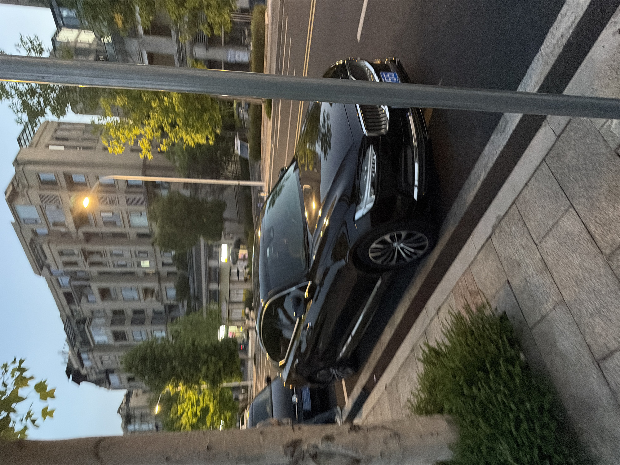 The image depicts a street scene in Jiangjin, China, possibly at dusk, with a black Volvo sedan parked on the right side of the road. The car is angled towards the viewer, showcasing its front grille and headlights. A light pole stands in the foreground, partially obscuring the car. Across the street, a row of multi-story buildings with modern architecture lines the horizon. Lush green trees and foliage are visible, some illuminated by streetlights, suggesting an urban park or residential area. The sidewalk in the foreground is paved with stone tiles and bordered by a low hedge. Another dark-colored car is parked to the left of the Volvo, with a portion of a person visible between the two vehicles. The lighting suggests it is either early morning or late evening.