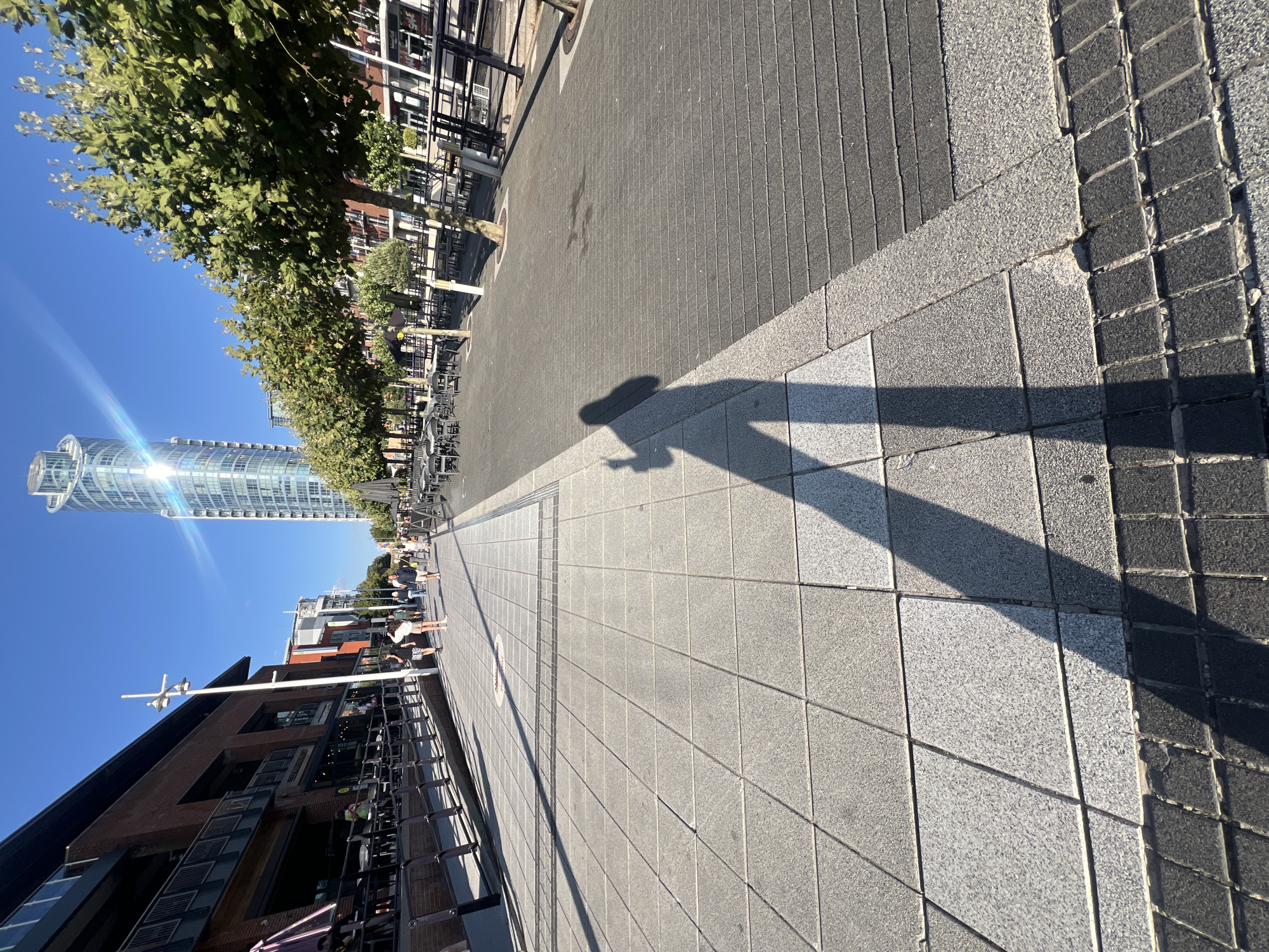 The image depicts a street scene in Portsmouth, United Kingdom, possibly near the Gunwharf Quays area. In the foreground, the shadow of a person holding a phone is cast on a tiled walkway. The walkway is made of large, grey square tiles with darker, rectangular tiles along the edge. Beyond the walkway, there is an area with outdoor seating for restaurants, indicated by tables and chairs. Several people are walking in the background. To the left, a brick building with multiple stories and balconies is visible, with awnings extending from some of the ground-floor establishments. To the right, a row of trees with green foliage lines the walkway. In the distance, a tall, modern skyscraper with a distinctive glass facade stands against a clear blue sky, with the sun glinting off its surface. The overall impression is that of a sunny day in a developed urban area.