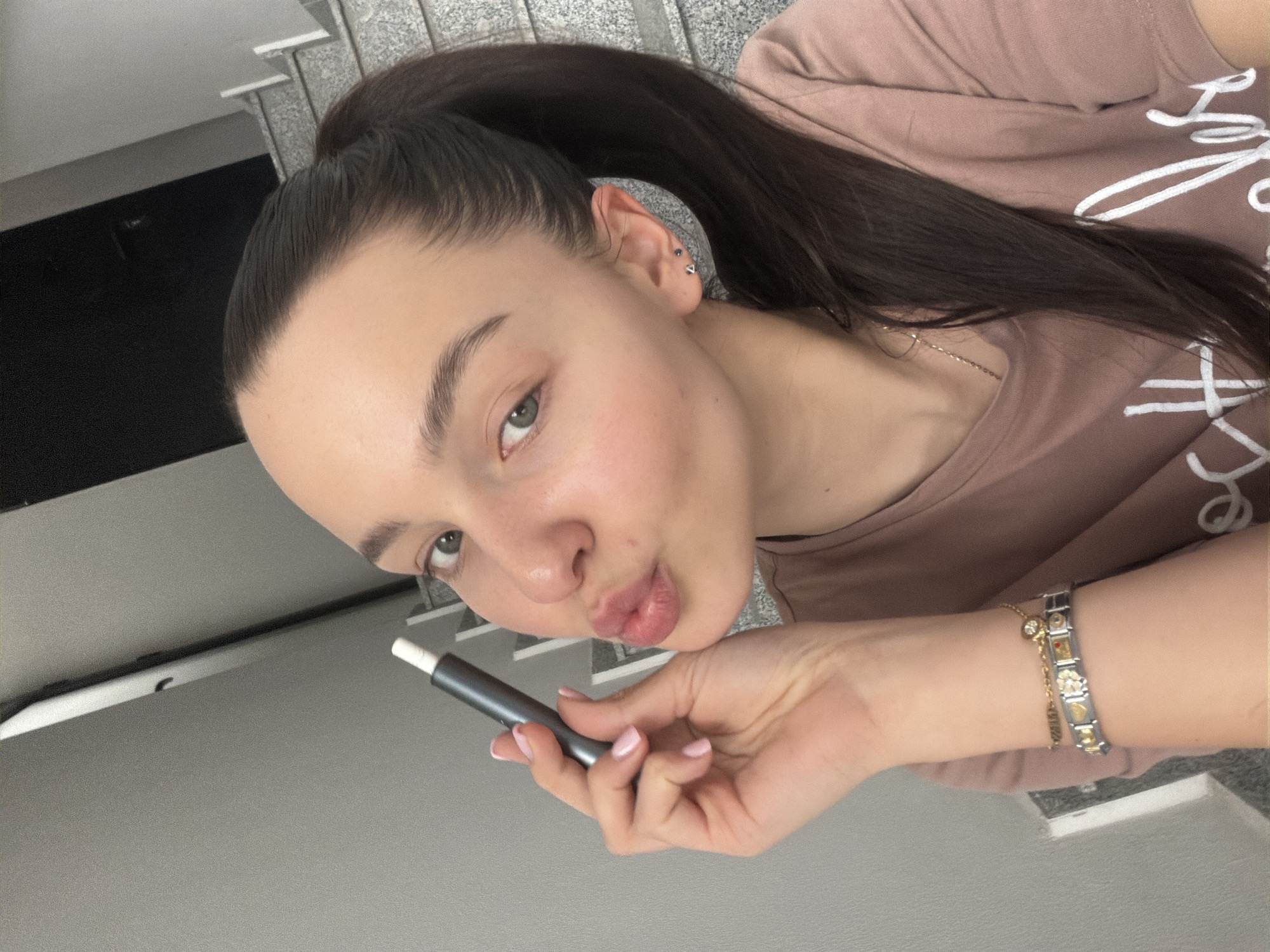 A woman is posing for a photo. She is holding a vaping device in her hand, with the tip illuminated. The woman has dark hair styled in a high ponytail. She is wearing a pink t-shirt. She is also wearing bracelets on her wrist. The background includes a gray staircase and a glimpse of a television. The location is likely indoors, and it is impossible to determine if this photo was taken in the city of Sofia, Bulgaria.