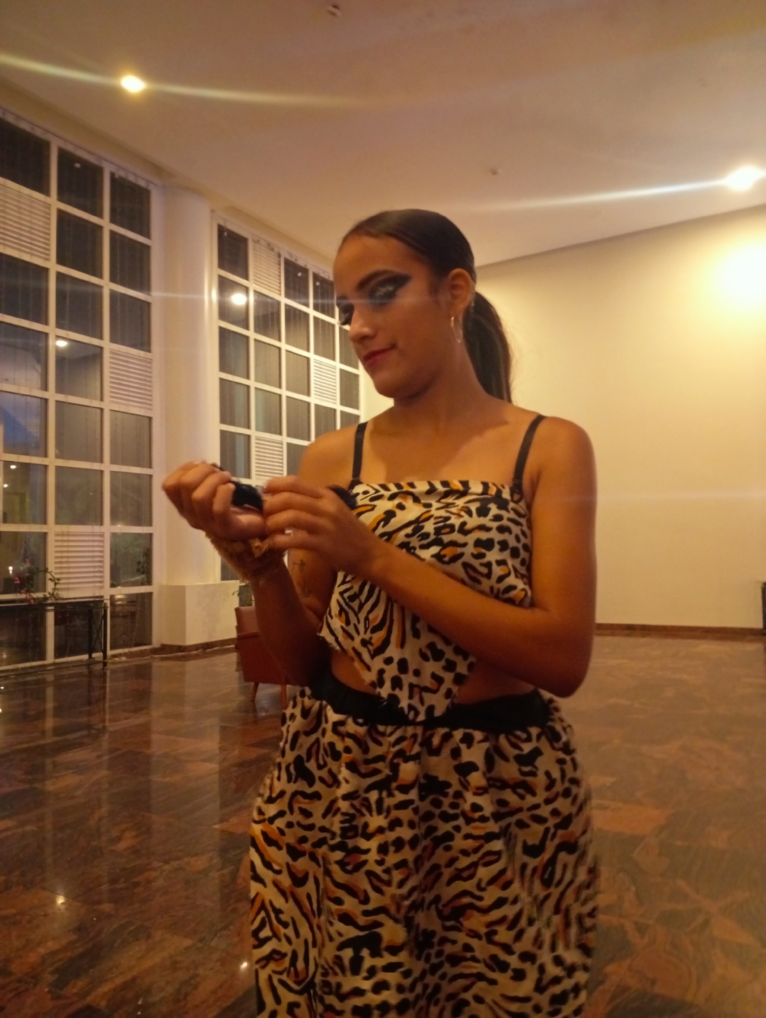 A woman is standing indoors in what appears to be a large room with a reflective, patterned floor. She is wearing a two-piece outfit with a black and tan animal print. The top has thin black straps and exposes her midriff, while the bottom appears to be a skirt or wide-legged pants with a black waistband. She has her dark hair pulled back into a ponytail and is wearing hoop earrings. Her makeup includes what appears to be dramatic eyeliner. She is looking down at something she is holding in her hands. The background includes large, gridded windows and a blank, light-colored wall. The overall lighting is soft and warm. The location is Pedro González, Venezuela.