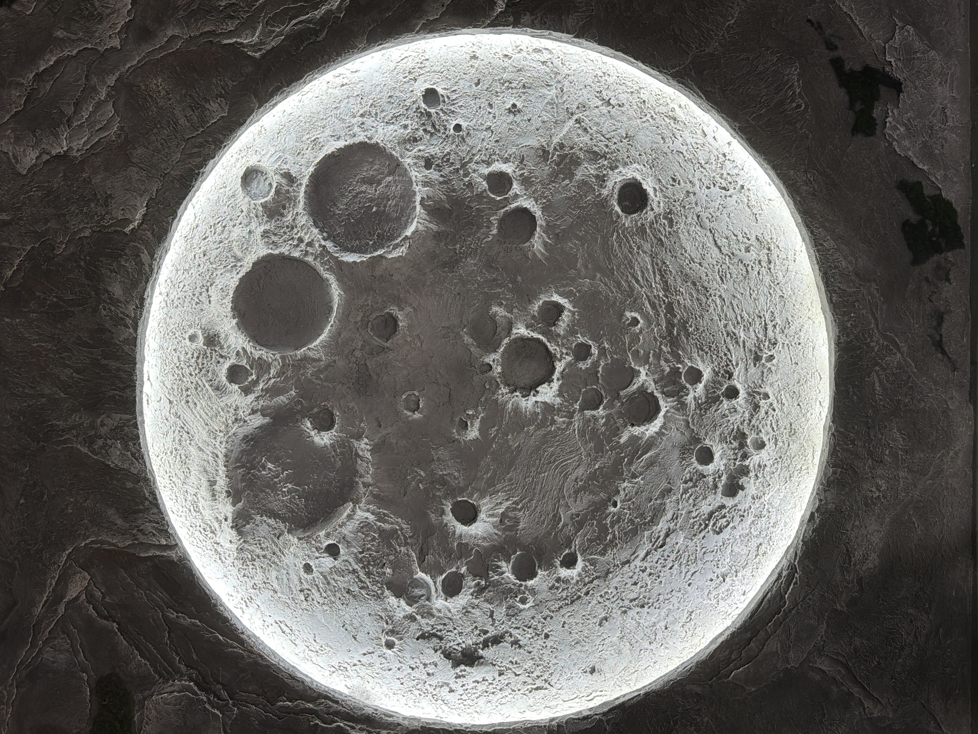 The image shows a close-up, detailed view of a large, illuminated model of the moon. The surface of the moon model is textured to represent craters and lunar terrain, with a bright light source behind it creating a halo effect. The model is set against a dark, textured background that appears to be a wall or ceiling with a rocky, uneven surface. There are no people or animals visible in the image. The lighting and texture suggest this could be part of an interior decoration, possibly in a themed room or a public space. There is no information within the image itself to indicate its location in Ulaanbaatar, Mongolia, or any specific city.