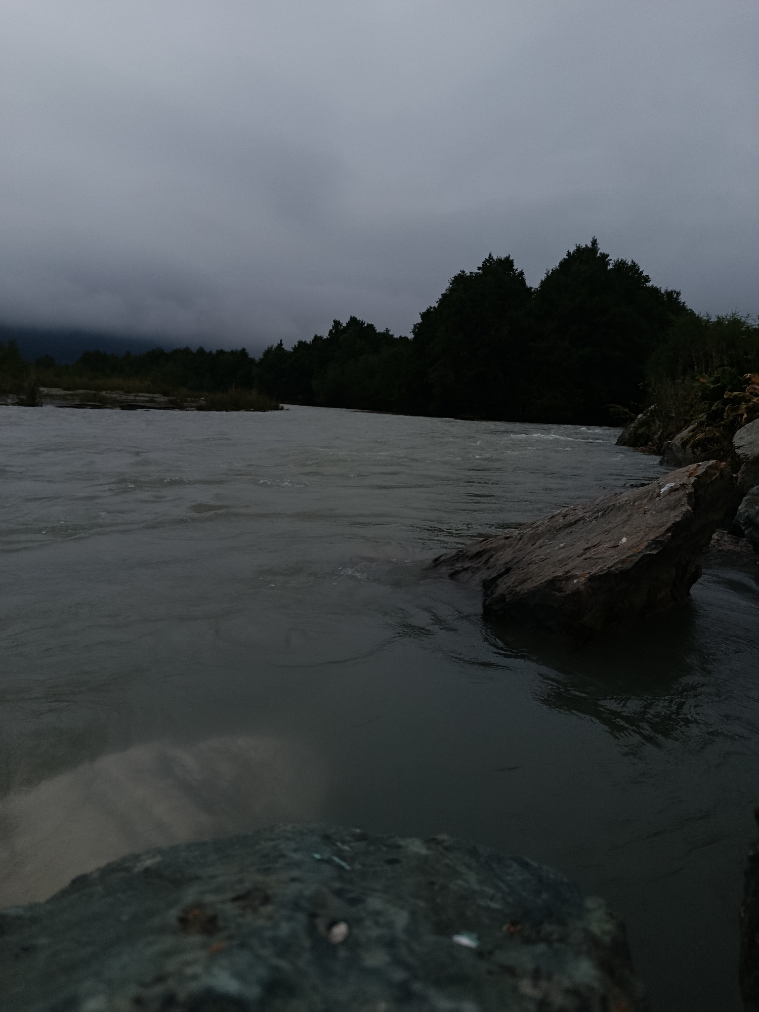 The image depicts a wide, fast-flowing river under a heavily overcast sky. The water is a murky grey, with visible ripples and currents. On the right bank, large, dark-colored rocks are partially submerged in the water, with some exposed above the surface. Beyond the rocks, dense, dark green trees form a thick forest that stretches along the riverbank. In the distance, the trees continue, and the land appears to rise slightly, meeting the low-hanging grey clouds that dominate the sky. The overall atmosphere is somber and moody, suggesting an overcast or twilight condition. The scene is natural, with no visible human presence or structures.