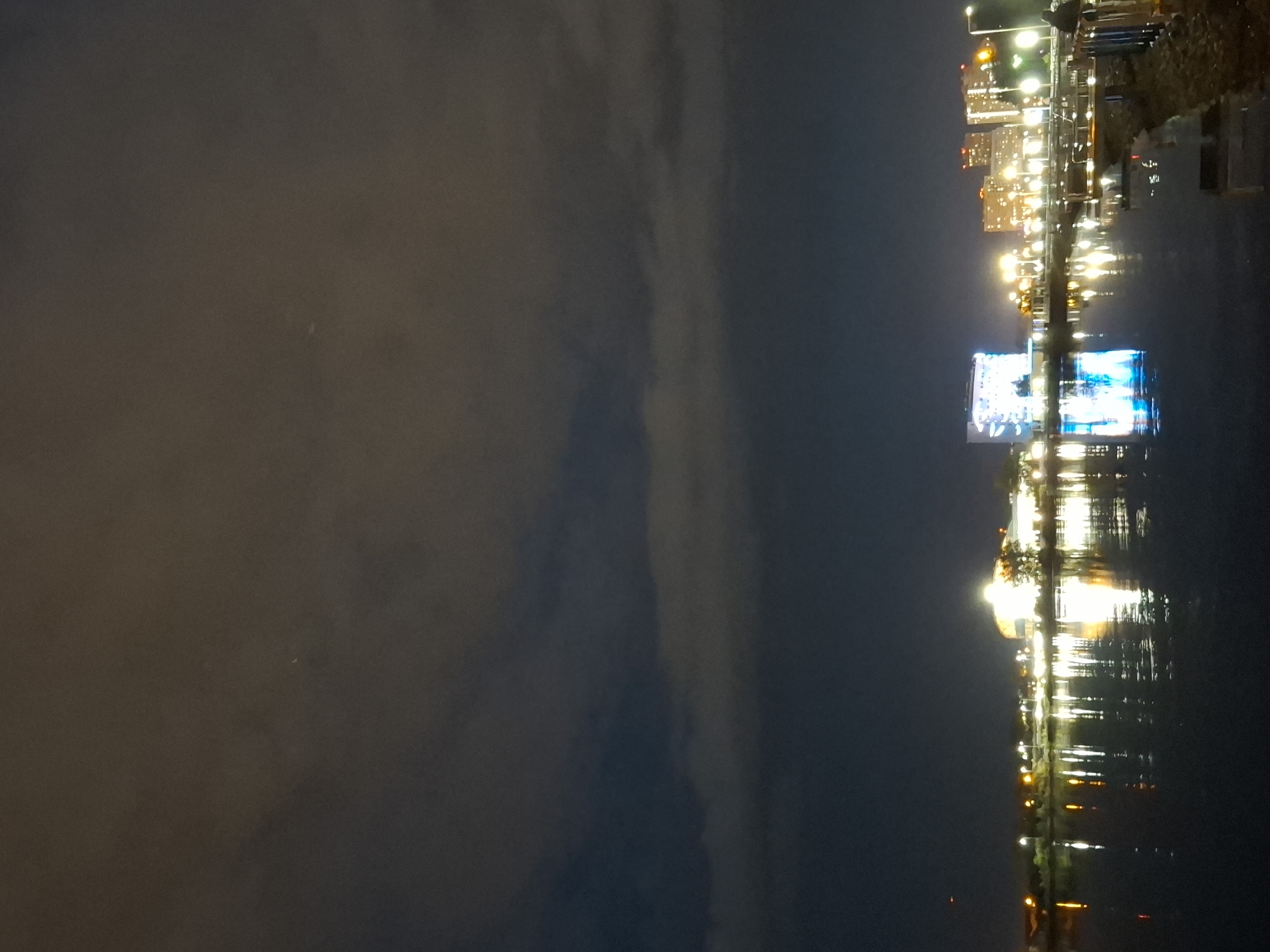An expansive nighttime aerial view showcases a body of water reflecting the illuminated skyline of Tyumen, Russia. The water's surface, dark and calm, mirrors the vibrant city lights, creating a shimmering effect. Buildings, both residential and commercial, are visible along the waterfront, with numerous windows aglow. A prominent structure in the mid-ground features a brightly lit facade displaying a colorful graphic. Streetlights punctuate the urban landscape, casting a warm, artificial light. The sky above is predominantly dark with patches of overcast clouds, lending a moody atmosphere to the scene. The overall impression is one of a bustling city at night, viewed from a tranquil vantage point.