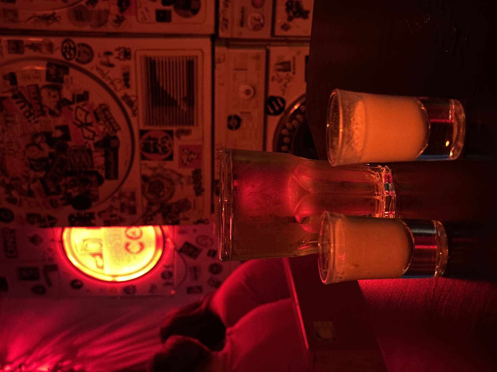 The scene is a dimly lit interior, bathed in predominantly red light, likely a bar or club setting. The focus is on a table on which there are two short glasses filled with a light-colored liquid, possibly a creamy or foamy beverage. Between them, a taller glass appears nearly empty. In the background, a wall is heavily decorated with stickers and small, framed items. A neon sign is visible, suggesting a business or establishment of some kind. A blurry figure, likely a person, is visible seated near the lower portion of the frame. While there is insufficient information to confirm the location in Saint Petersburg, Russia, as requested, the photograph depicts a nighttime gathering place with a beverage-centric focus.