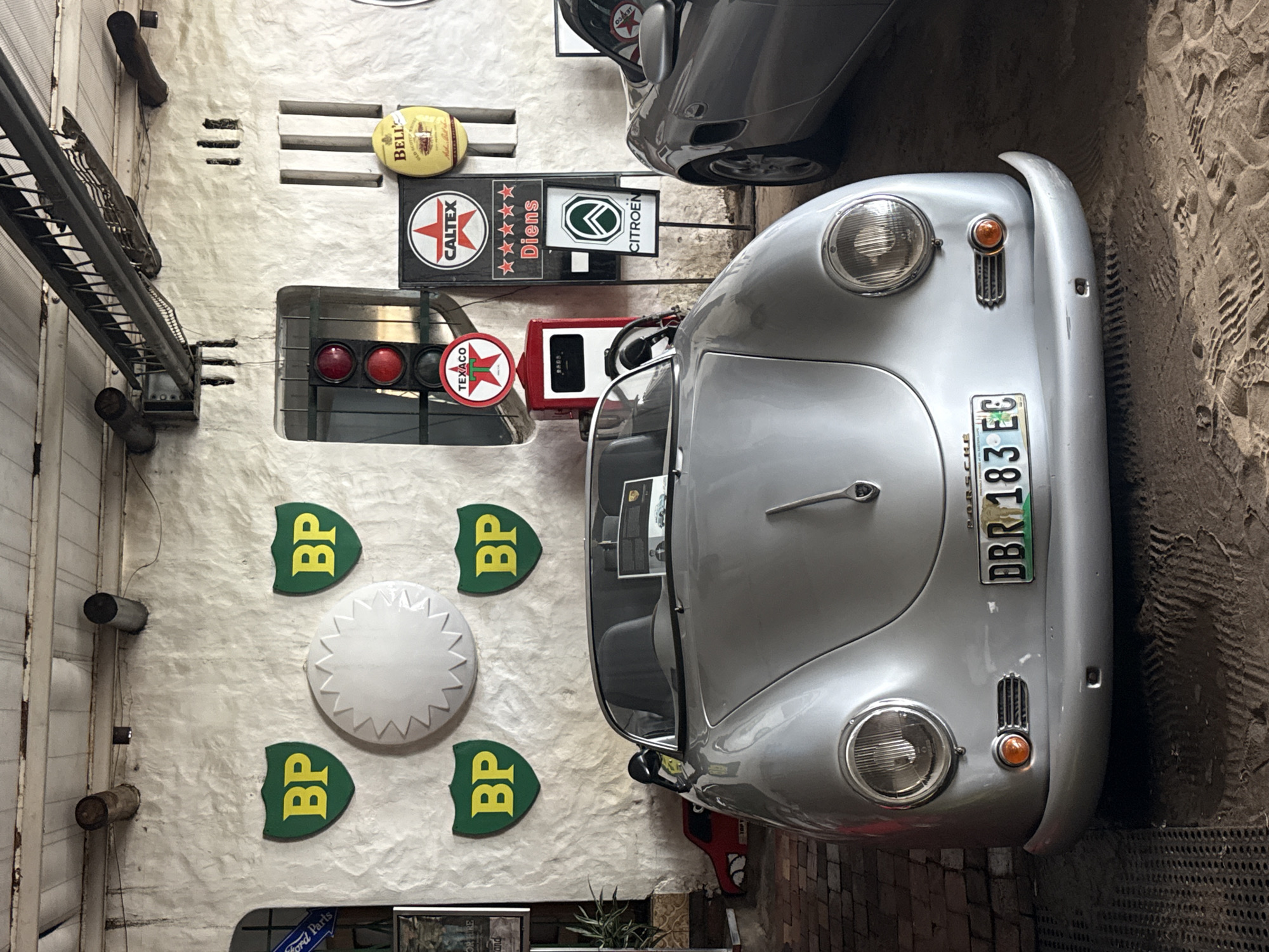 The image captures a scene that seems to blend automotive aesthetics with an industrial setting. A silver Porsche is prominently featured, parked near a collection of vintage gas and oil company signs mounted on a textured white wall. The signs include logos for BP, Caltex, Texaco, Citroen, and a Bell product. A traffic light and a vintage gas pump add to the automotive theme. An overhead structure, possibly a metal ladder or scaffolding, runs along the left side of the frame. Below the signs, near the bottom, a partial view of a "Ford Parts" sign is visible, suggesting a potential automotive repair shop or garage. The ground near the Porsche appears to be sand or dirt with visible tire tracks. The scene is in Kraaifontein, South Africa.