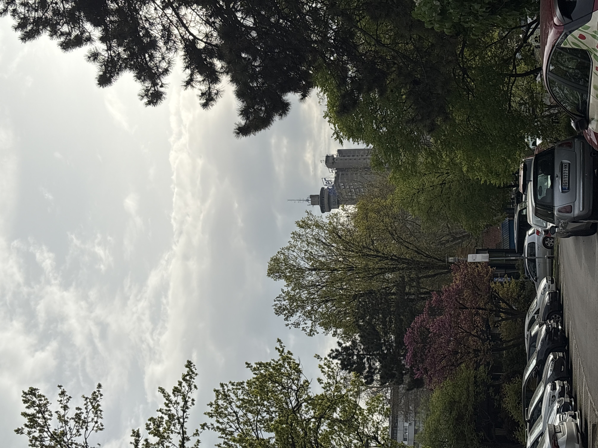 The image shows a view of Belgrade, Serbia. In the distance, a telecommunications tower and several apartment buildings are visible. Trees line the street, some with pink blossoms. Cars are parked along the side of the street. The sky is cloudy.