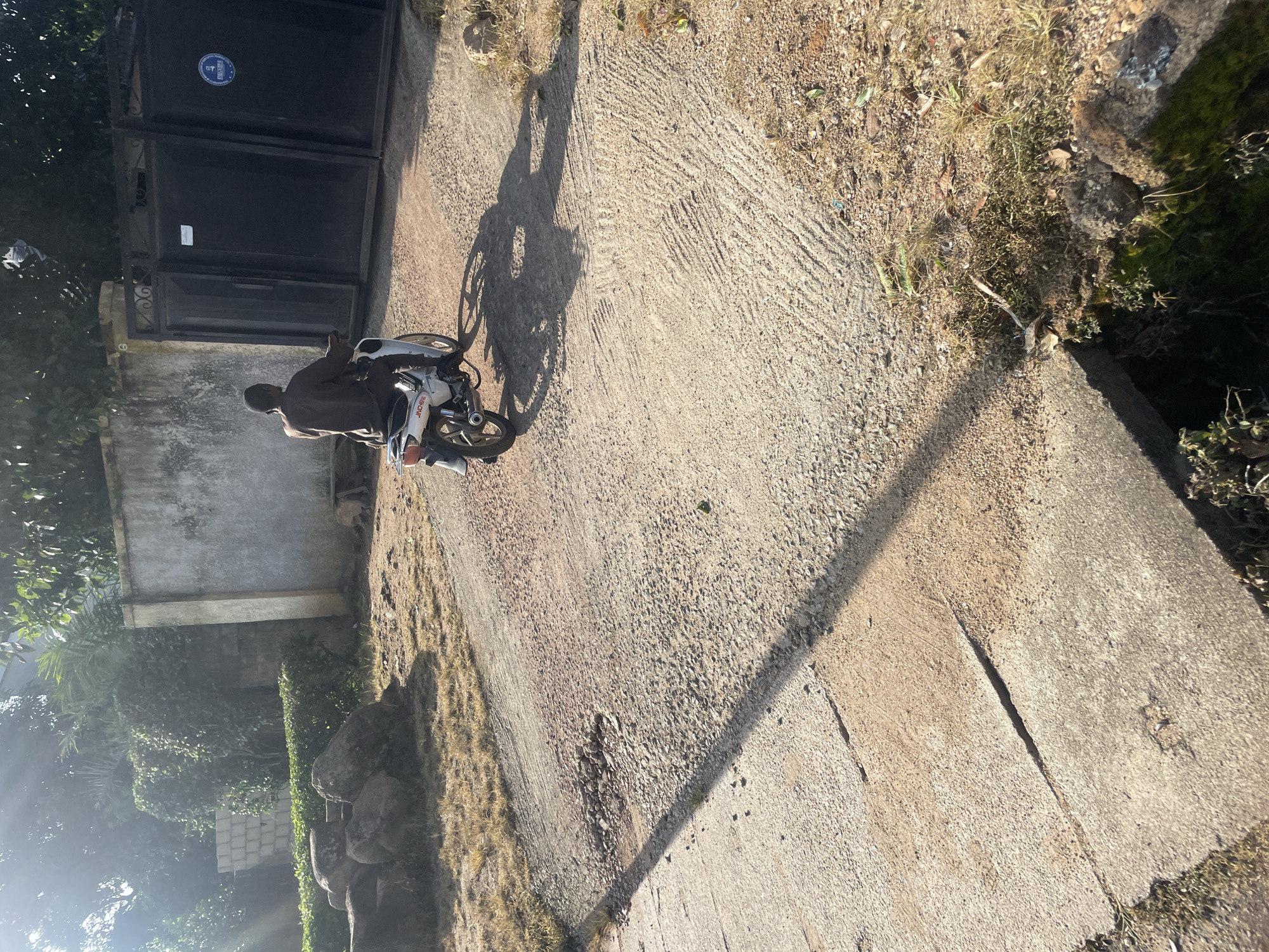 A man rides a white motorcycle down a rough, uneven concrete and dirt path in Jos, Nigeria. The man wears a dark brown tunic and pants. The motorcycle is heading towards a large, black metal gate. The gate is flanked by a concrete wall on the left and a smaller, darker gate on the right. Sunlight streams in from the left, casting long shadows across the path. Bushes and trees are visible behind the wall. There are rocks and dry grass scattered on the ground. The scene appears to be an outdoor area, possibly a driveway or entrance to a property.