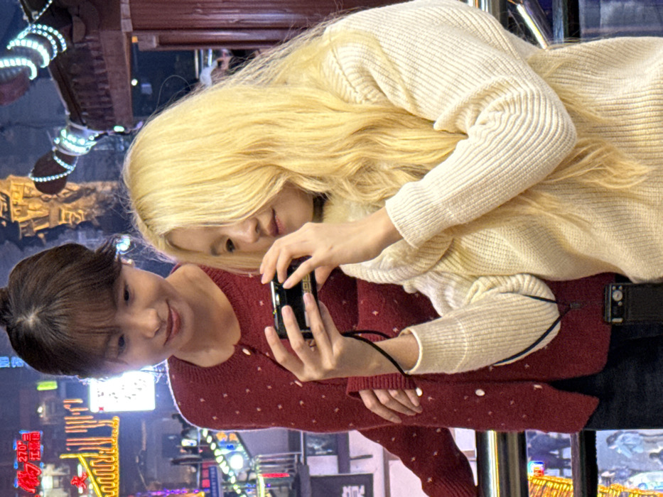 This nighttime image depicts two East Asian females in an illuminated urban environment within Yuzhong District, China. The foreground subject, characterized by long, wavy blonde hair and a cream-colored ribbed turtleneck sweater, holds a black compact digital camera with both hands, aiming it forward as if capturing an image. Her gaze is directed towards the camera. Behind and slightly to her left, a second female, with dark brown hair styled with bangs and wearing a red cardigan with white polka dots, stands observing. Her head is tilted downwards towards the camera held by the foreground subject, and her right hand rests on the foreground subject's back. The background is brightly lit with various commercial and decorative lights, including prominent yellow and red neon signs, and blue-lit outlines of traditional architectural elements, specifically curved eaves. Blurred figures of other individuals are visible in the distant background, indicating a populated area. A dark horizontal barrier runs across the lower middle section of the image.