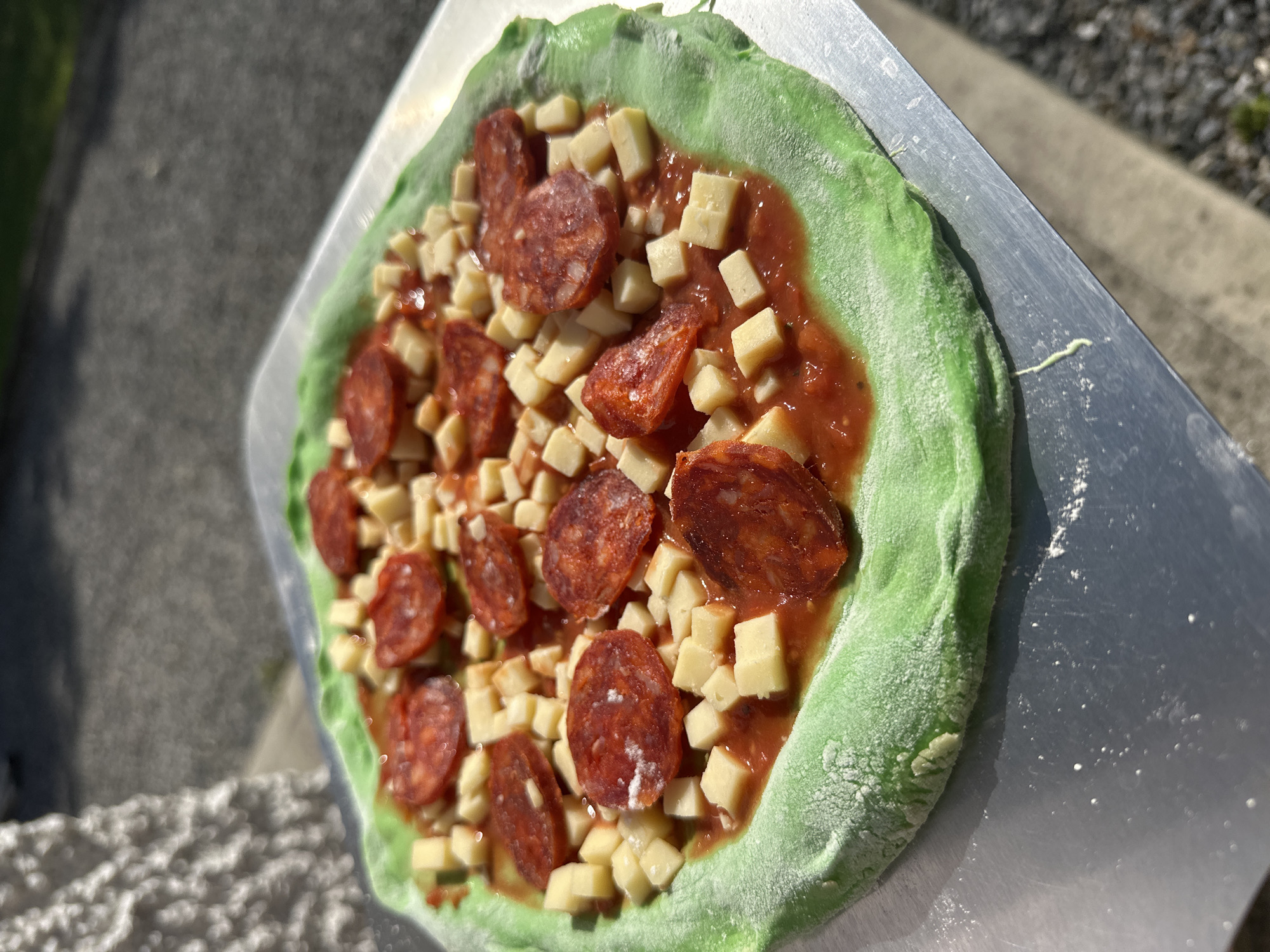 A close-up, overhead shot shows a green-crusted pizza topped with diced cheese, tomato sauce, and slices of pepperoni. The pizza rests on a metal peel, likely ready for baking. The background is blurred, suggesting an outdoor setting with sunlight dappling on a textured surface, possibly a sidewalk or stone wall. The specific location in Moortown, Ireland, is not discernible from the visual information provided.