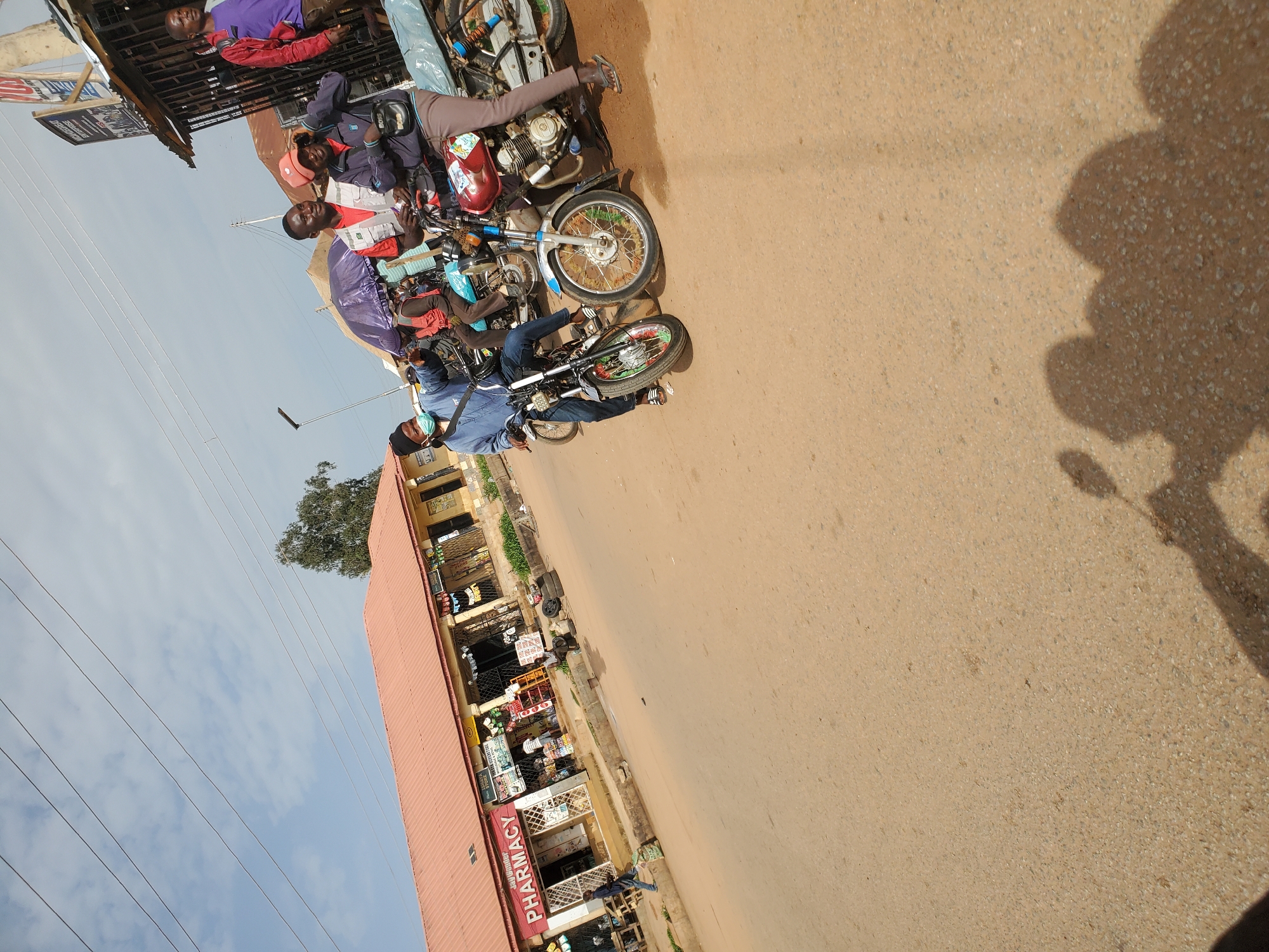 In Bukuru, Nigeria, a street scene unfolds under a cloudy sky. A row of businesses lines the left side of a paved road, including a "PHARMACY" store with a red awning and a smaller store with various goods displayed outside. Several people are present, some standing and interacting, while others are seated on motorcycles. In the foreground, a person wearing a blue shirt and a dark cap rides a motorcycle, with another motorcycle to their right. Further back, a group of men are gathered, some holding what appear to be phones. The overall environment suggests a bustling commercial area with motorcycle transportation being prominent.
