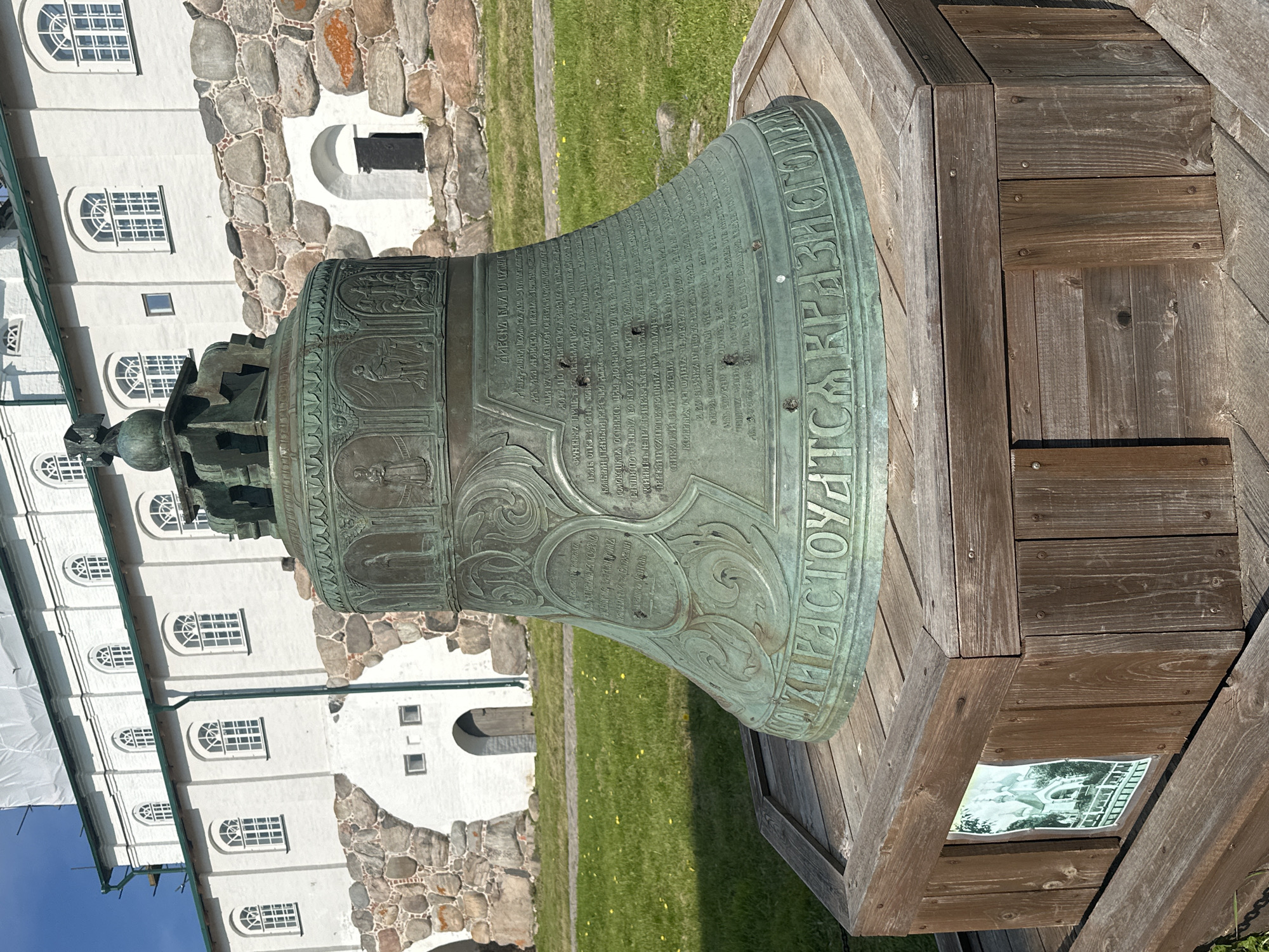 A large, ornate bronze bell sits on a wooden pedestal in front of a white stone building with arched windows. The bell is covered in intricate carvings and inscriptions. The building appears to be a historical or religious site, possibly a monastery given the location. The sky is blue and the ground is covered in green grass. The overall scene suggests a historical landmark in Solovetskiy, Russia.