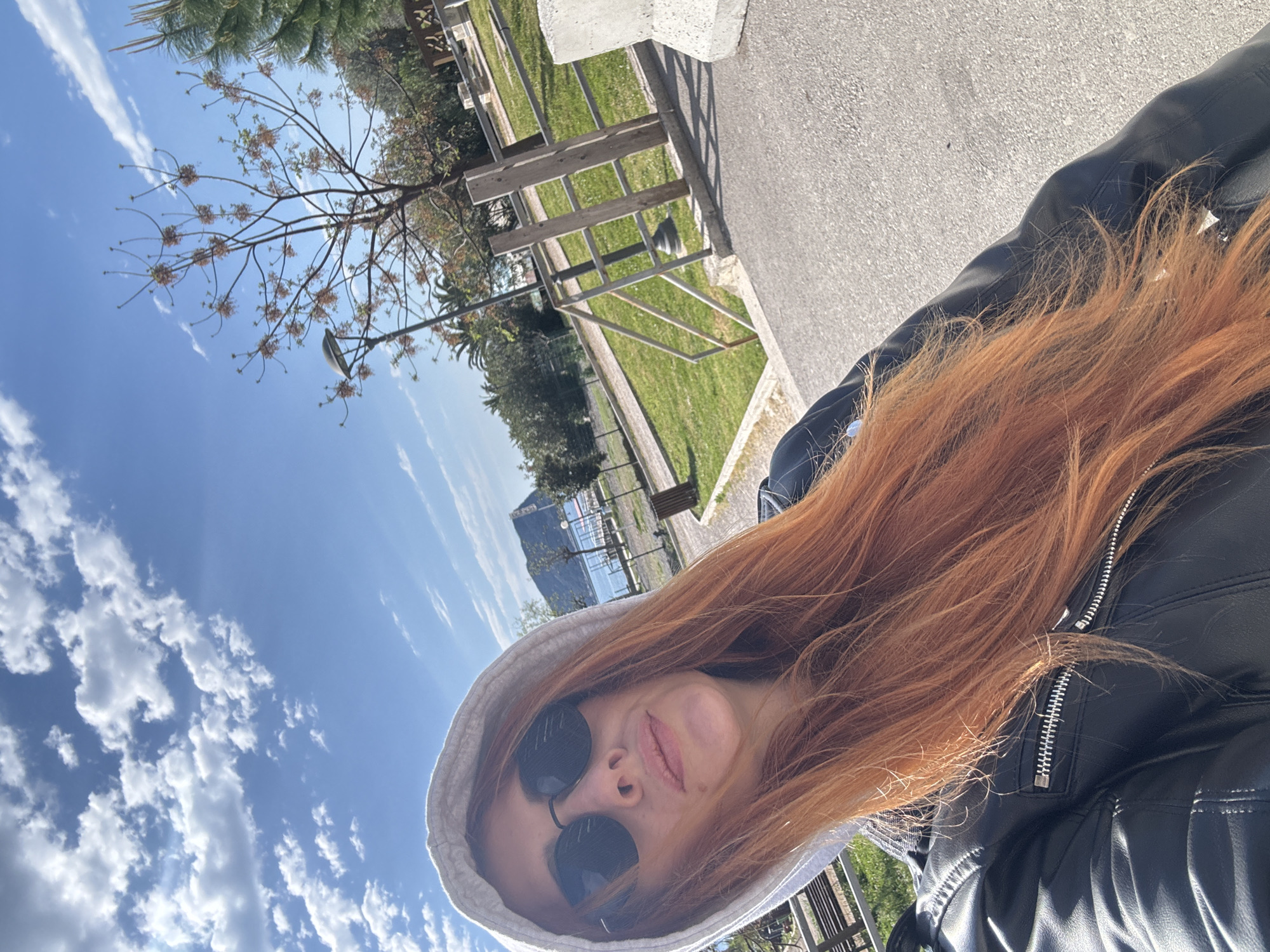 A woman with red hair and sunglasses is wearing a white hoodie and a black leather jacket. She is standing outside on a sunny day with a blue sky and some clouds. Behind her is a tree, a grassy area, a bench, a railing, and a body of water. The location appears to be a park or walkway near the sea, which can be associated with Budva, Montenegro.