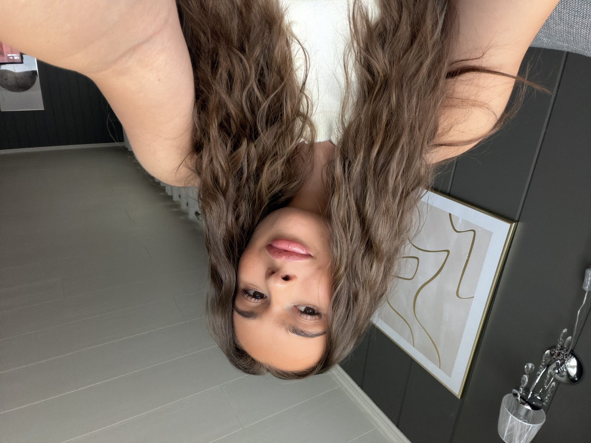 The image shows a young woman with long, wavy brown hair, looking directly at the camera. She is wearing a white top. The background appears to be an indoor setting with a dark grey wall, a framed piece of art with abstract gold lines on a light background, and a light fixture on the wall. The ceiling is visible and has a slatted design. The scene is well-lit. There is no indication of the location being in the city of Harstad, Norway.
