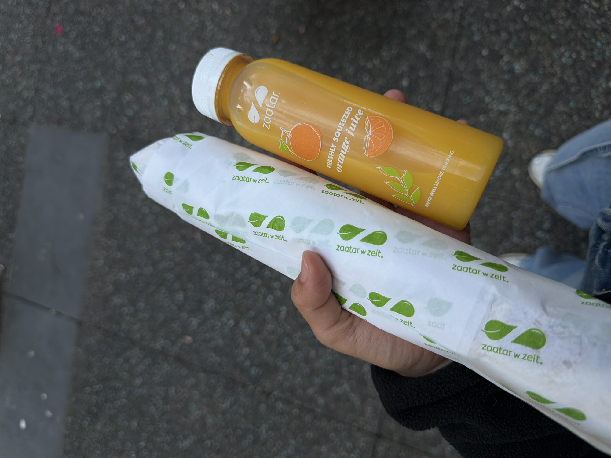 A person's hand is holding a long, white paper-wrapped food item and a bottle of orange juice. The food item has a repeating pattern of green leaves and the text "zaatar w zeit". The orange juice bottle is transparent, filled with orange liquid, and has a white cap. The label on the bottle features the brand name "zaatar" at the top, with images of an orange and an orange slice below. Text on the label reads "FRESHLY SQUEEZED orange juice" and "SHAKE WELL BEFORE DRINKING". The background appears to be a sidewalk or paved ground with a mottled texture. The overall impression is of a casual, outdoor setting, potentially in a city like Vancouver, Canada, given the urban pavement. The image is taken from a slightly overhead perspective, focusing on the items in the person's hand.