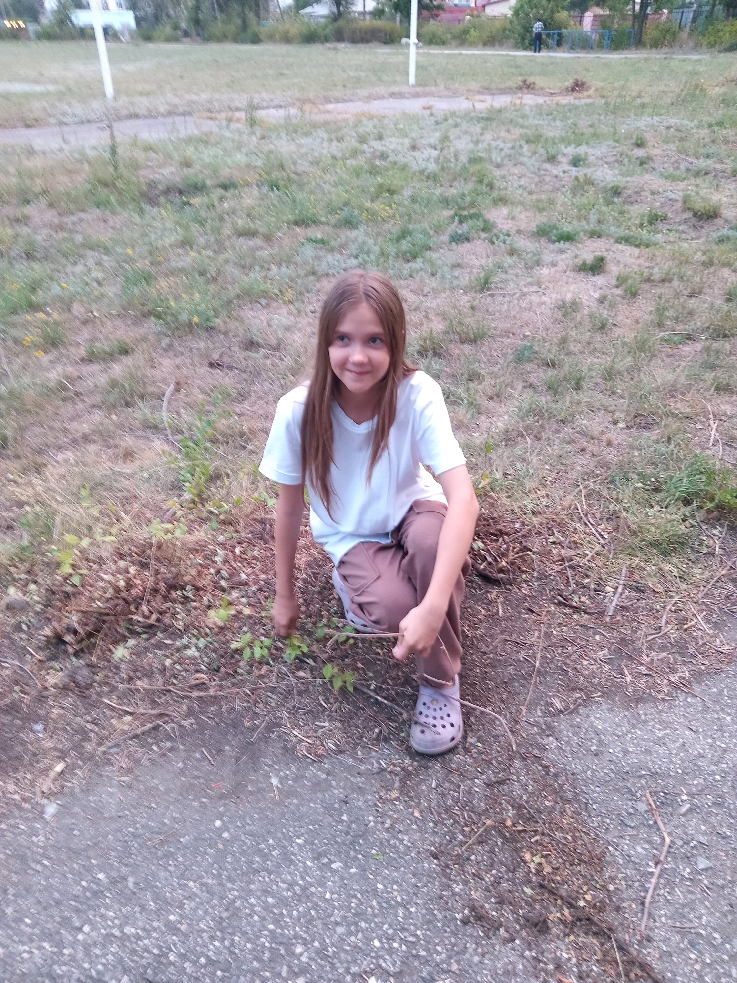 A young girl with long, light brown hair is crouched down on a patch of dry grass and dirt, looking towards the camera with a slight smile. She is wearing a white t-shirt and brown pants. Her left foot is visible, clad in a light purple Croc-style shoe. She is holding a small stick or twig in her hands, appearing to be playing with it. The background shows a grassy field with some scattered debris and a paved area in the foreground. In the distance, there are trees and buildings, suggesting a semi-urban or suburban setting. The lighting and general appearance of the vegetation suggest it might be a warm day, possibly late afternoon or early evening. There is no visible text in the image. The setting appears to be Rudny, Kazakhstan.