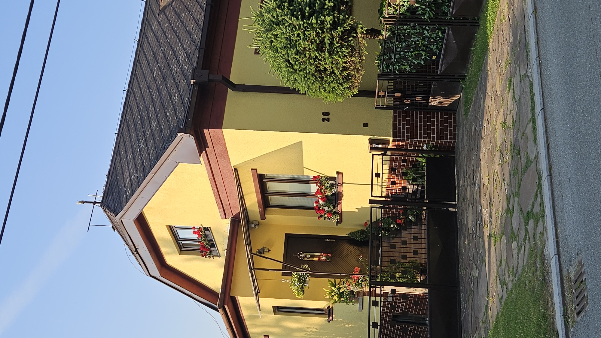 The image shows a bright yellow house with a dark shingled roof. The house has at least two windows, both adorned with flower boxes filled with red and white flowers. The front door is dark wood with decorative panels and a hanging lamp above it. A black metal gate encloses the property, which features a paved driveway made of irregularly shaped stones and some grassy areas. There is a small, rounded evergreen tree to the right of the house. The number "26" is visible on the exterior wall of the house. The sky is clear and blue, suggesting a sunny day. The scene appears to be a residential street in the city of Prešov, Slovakia.