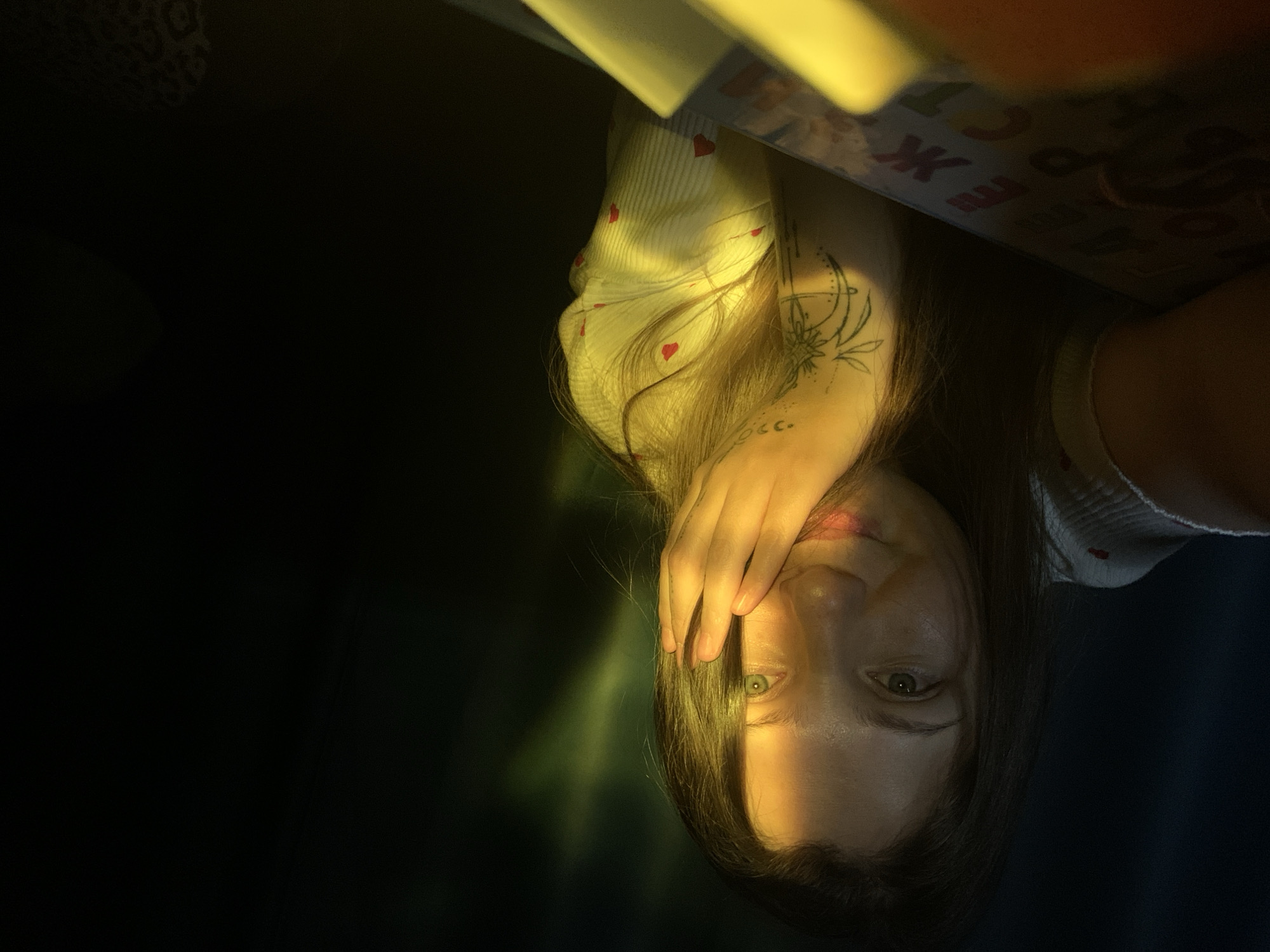 A woman is depicted in an upside-down close-up shot. She is covering part of her face with her hand. Her hair is dark and falls across her face. She has light skin and visible tattoos on her arm. She is wearing a light-colored shirt with red heart patterns. The background is mostly dark, with some light emanating from above, illuminating the woman and some objects above her. There is no clear evidence to suggest the location is Chaikovsky, Russia.