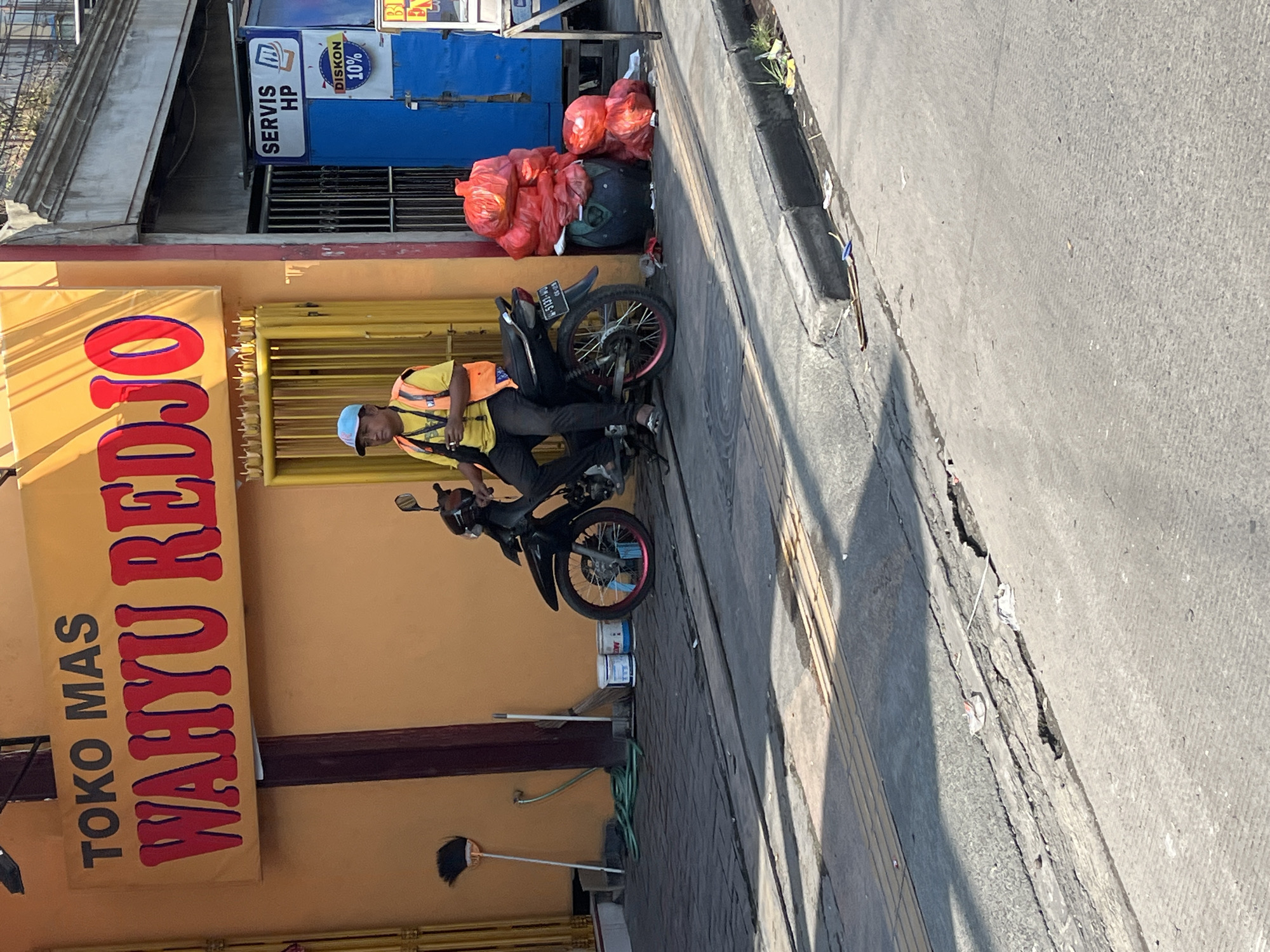 A medium shot shows a man in a blue cap and an orange safety vest sitting on a black motorcycle. He is on a sidewalk next to a building with a yellow facade and a sign that reads "TOKO MAS WAHYU REDJO". To the right of the building, there is a blue structure with a "SERVIS HP" sign and a smaller sign advertising a "DISKON 10%". A pile of red plastic bags is next to the blue structure. The man appears to be looking to his left. The sidewalk is paved with dark grey bricks, and a cracked concrete road is in the foreground. There are several brooms, buckets, and a green hose on the sidewalk behind the motorcycle. The scene is outdoors and appears to be in a commercial area.