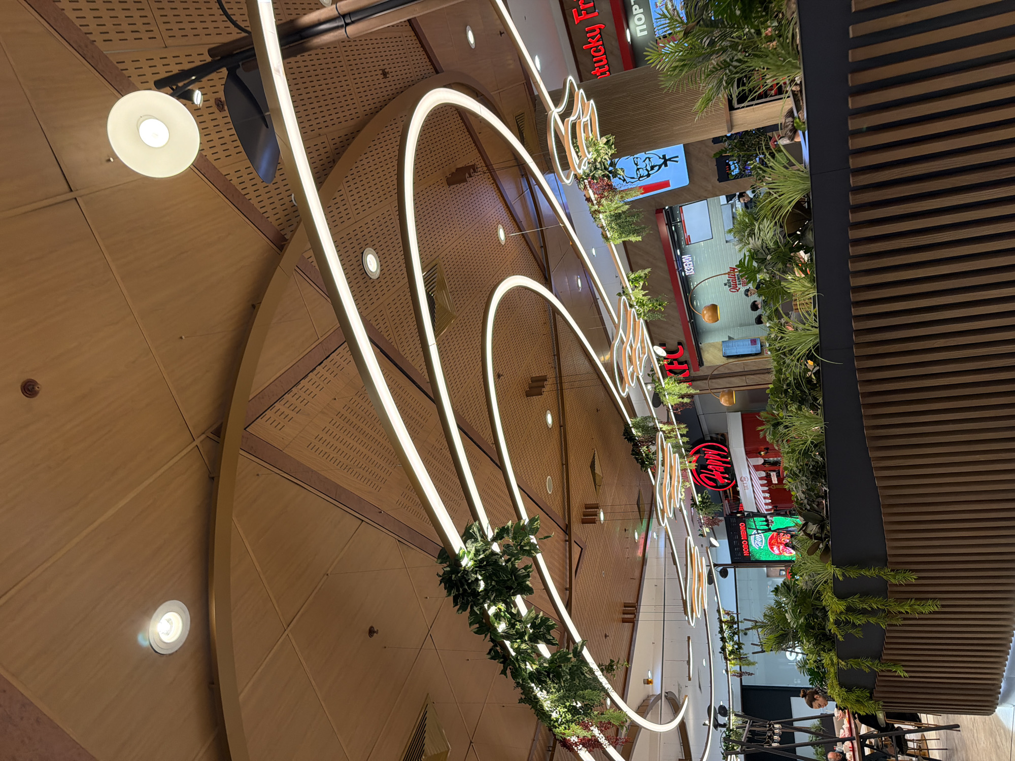 This image captures the interior of a modern shopping mall or food court in Sofia, Bulgaria, characterized by a clean and contemporary design. The perspective is looking upwards and across, highlighting the architectural elements.
The most striking features are the large, curvilinear light fixtures that span across the ceiling or upper levels, emitting a bright, continuous glow. These light structures are partially adorned with artificial greenery, adding a touch of natural aesthetic to the urban indoor setting. The ceiling and upper walls are clad in light-colored wooden panels, some featuring a perforated pattern. Spotlights are also integrated into the ceiling, providing additional illumination.
In the background, several commercial establishments are visible. Text on signs indicates food and beverage outlets, including a prominent "Kentucky Fried Chicken" (partially visible as "Kentucky Fr") with its distinct "KFC" logo. Another food stall displays the word "Quality" on its signage, and a red sign that appears to say "MAMY" or "НАПИ" (Napi) is also seen. A digital display screen shows advertisements, including text that partially reads "OHEN OOCN" or similar in Cyrillic, along with images of food. Another sign in Cyrillic reads "ПОРТ" (Port).
A few individuals are present, mainly in the background near the shops. One person, a woman with her hair tied back, is clearly visible behind a counter or stall in the lower right section of the image, appearing to be a staff member. The environment is well-lit by artificial lights, suggesting daytime hours of operation for the commercial space. The overall atmosphere is modern, bright, and bustling, typical of a shopping center or food court.
