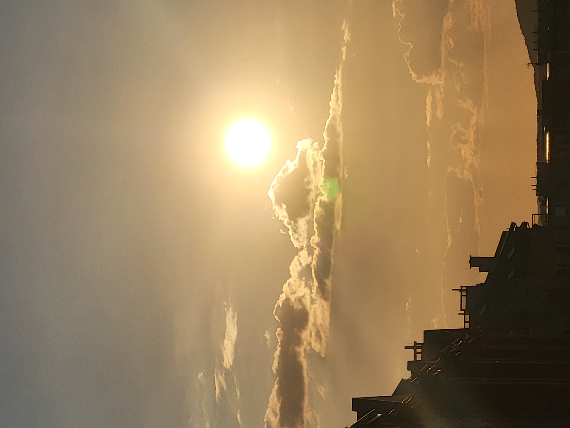 An outdoor vertical, eye-level, medium shot captures the sun setting in a sky filled with clouds over a city skyline. The sun is a bright white orb centered in the upper right quadrant of the image, surrounded by a soft yellow glow. Various clouds are scattered throughout the sky, some illuminated by the sun, others casting shadows. The lower portion of the image is dominated by the silhouettes of buildings with pitched roofs and chimneys. The overall color palette is warm, with hues of yellow, orange, and a soft greyish-blue in the upper sky. The scene appears to be a calm evening in an urban environment.