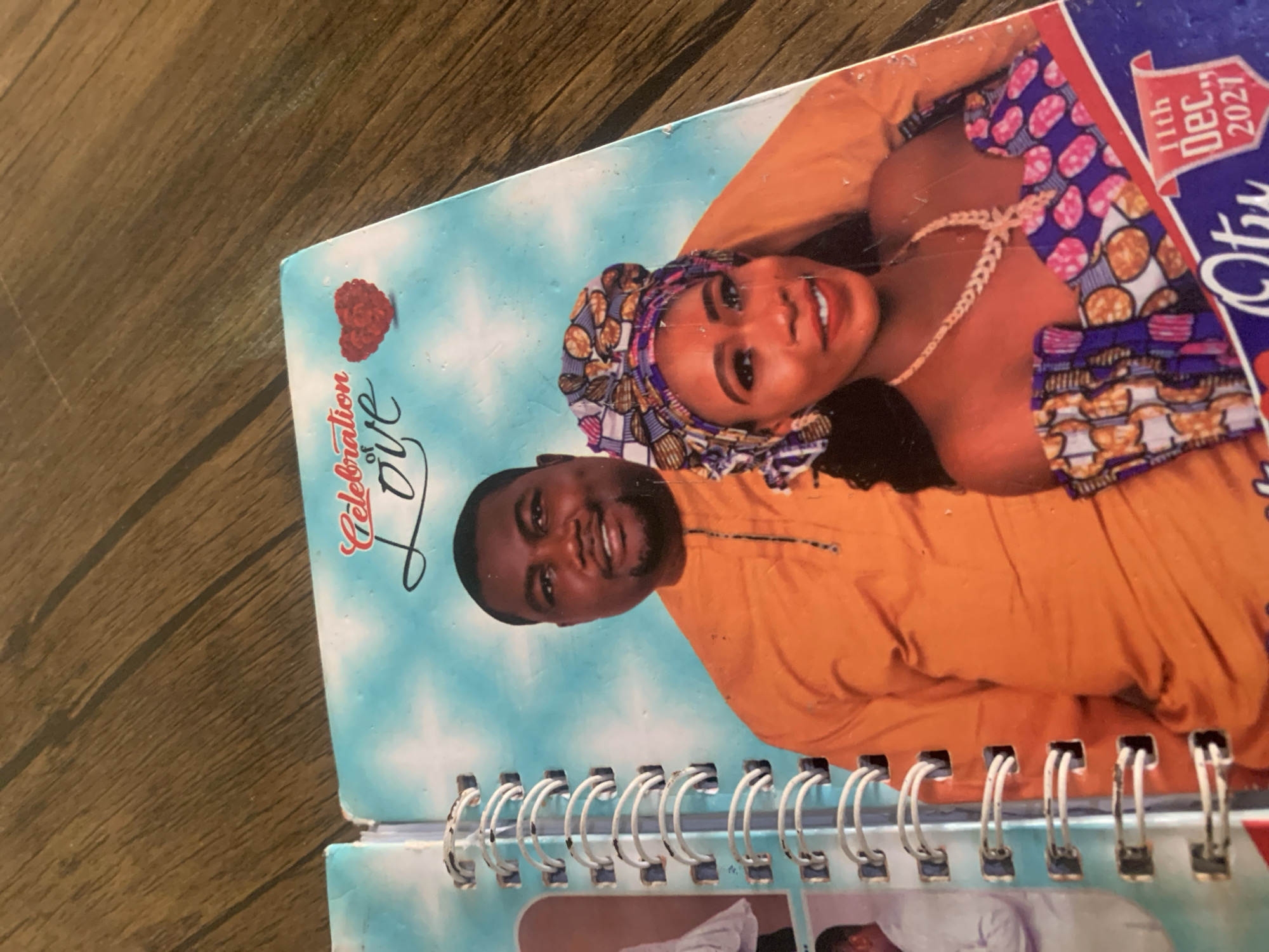 A close-up, eye-level shot shows a page from a spiral-bound photo album or booklet lying on a wooden surface. The page features a colorful illustration of a sky with stylized clouds, overlaid with text that reads "Celebration of Love" in a cursive font. A red, textured heart shape is positioned near the text.
Dominating the page are two individuals, a man and a woman, who appear to be a couple. The man, on the left, is wearing an orange traditional garment with a simple neckline. He has dark skin, a shaved head, and is smiling towards the camera. The woman, on the right, is adorned in a vibrant, patterned headwrap and a matching off-the-shoulder top. She has dark skin and is also smiling. She is wearing a prominent, multi-strand beaded necklace with a pendant.
To the left of the main couple, smaller inset photos are visible, showing other individuals. The left edge of the page reveals the white spiral binding mechanism.
In the lower right corner of the page, a red banner with white text announces "11th Dec., 2021" and part of a name, "Otu" is visible in large, white lettering below. The overall impression is one of a celebratory or commemorative occasion. The wooden surface appears to be a table or countertop with a visible grain pattern. The image does not contain information about the city of Jauro Yinu, Nigeria.