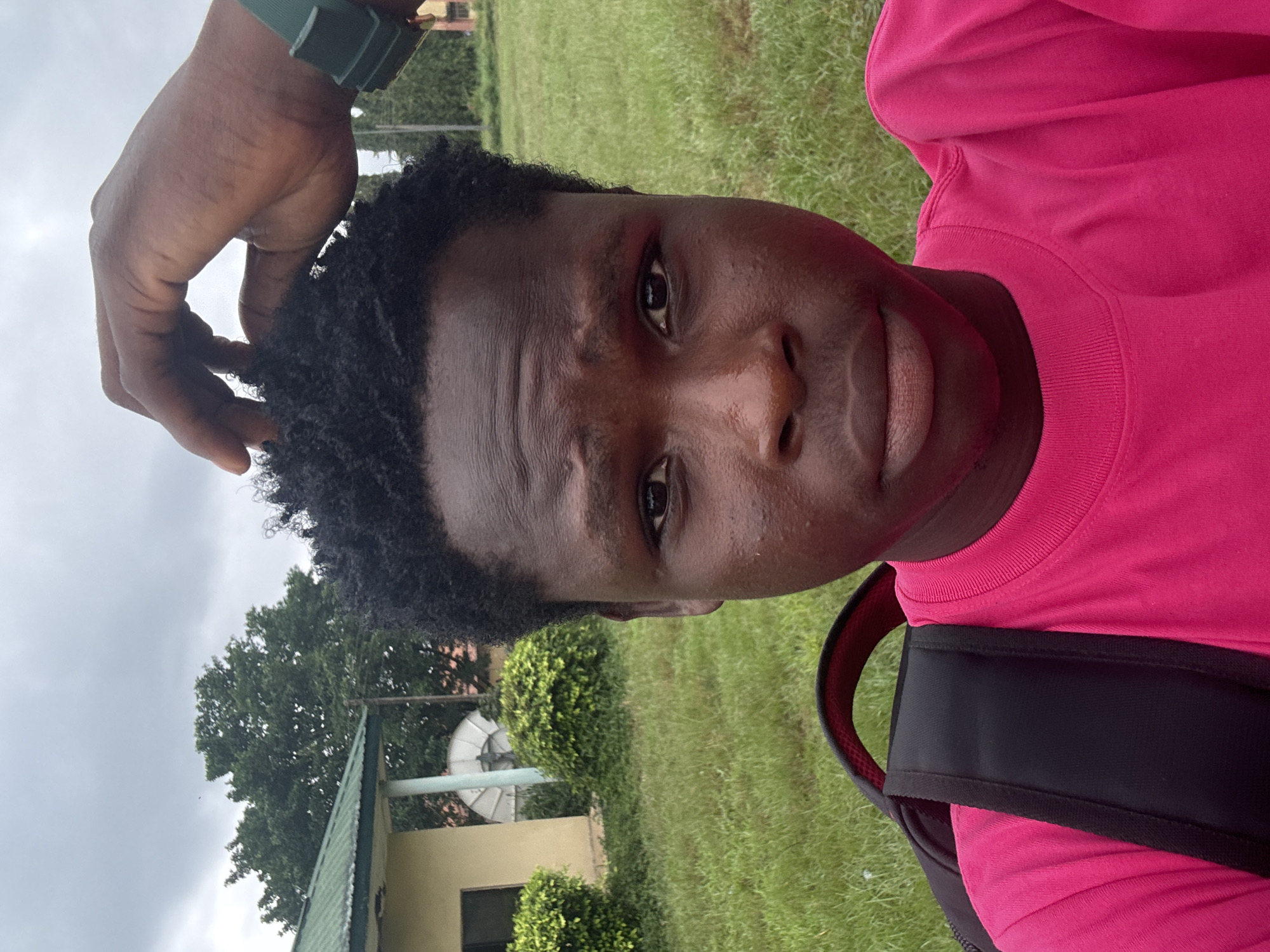 A close-up, outdoor, selfie-style photograph shows a young Black person, possibly male, with dark, short, curly hair. The person is wearing a bright pink t-shirt and a black backpack. Their right hand is visible touching their hair. The background features a grassy area with a building and trees, suggesting a rural or suburban setting in Ihiagwa, Nigeria. The sky is overcast. The overall lighting is diffused, typical of a cloudy day. The focus is on the individual, with the background slightly blurred.