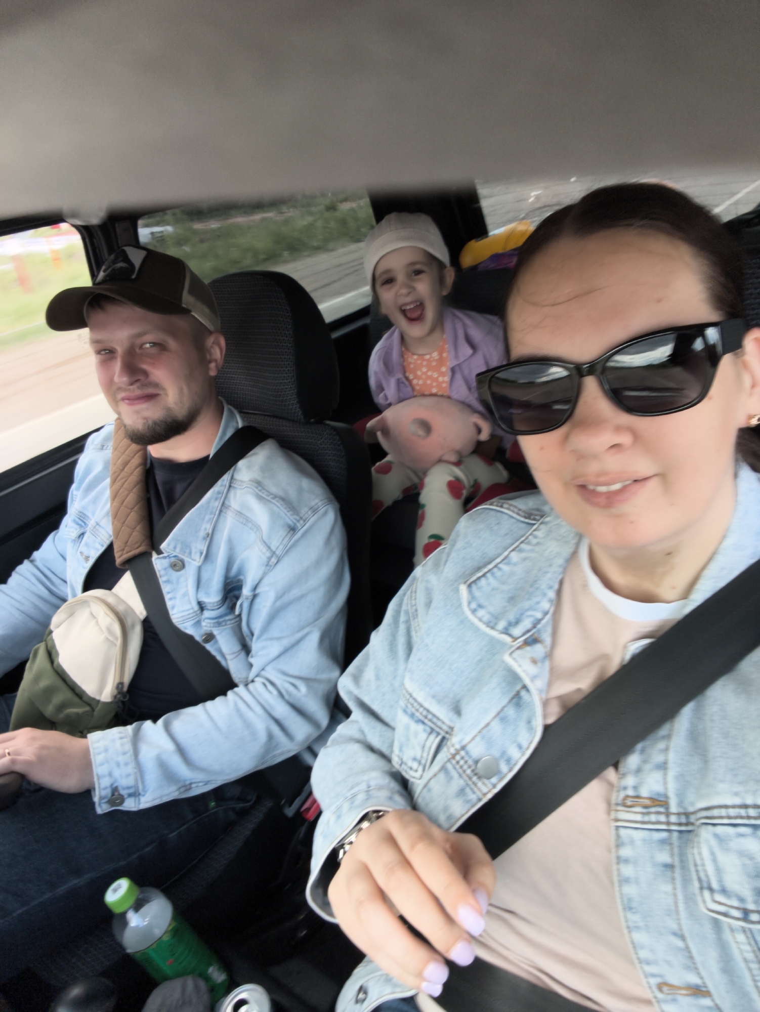 The interior of a car is depicted in the photograph, with three occupants. In the driver's seat, a woman with sunglasses is visible, wearing a denim jacket and a seatbelt. A man in a denim jacket and a baseball cap is in the passenger seat, also wearing a seatbelt and a shoulder bag. A child with a white hat, wearing a purple shirt and strawberry-patterned pants and holding a pink stuffed toy, is in the back seat. Bottles and cans are visible on the floor of the car. The location is Chaikovsky, Russia, based on information available.