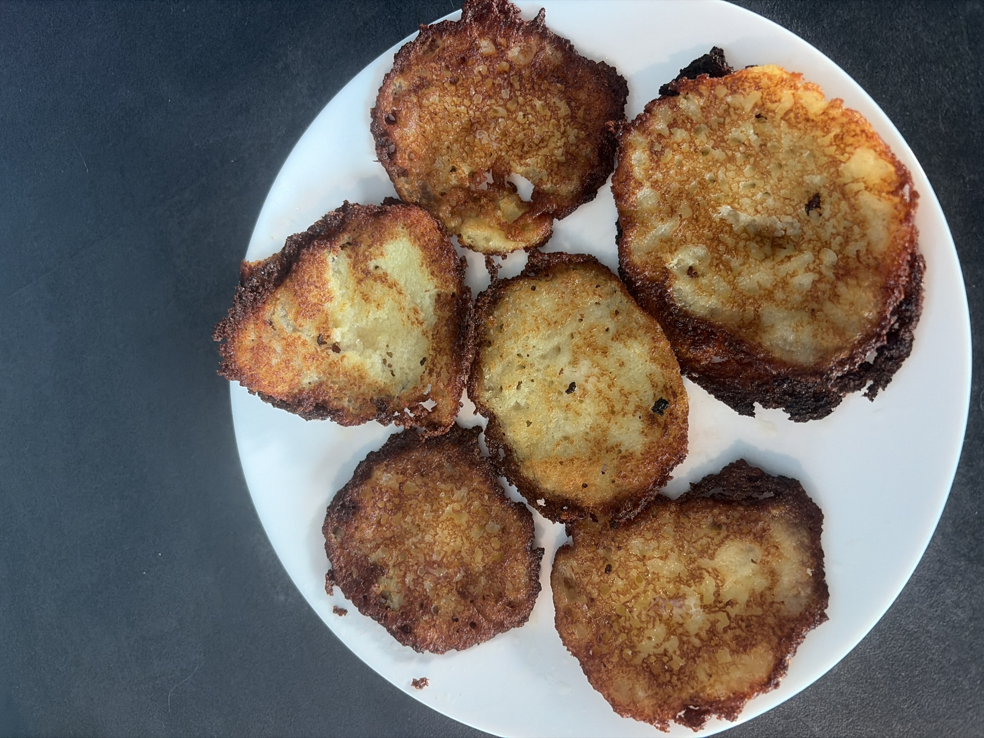 The image displays a close-up, overhead view of six golden-brown, pan-fried potato pancakes arranged on a plain white, round plate. The pancakes vary slightly in size and shape, with irregular, crispy edges that range in color from deep golden to dark brown, indicating a good char from frying. The surfaces of the pancakes show the texture of grated potato, some areas appearing lighter yellow and softer, while others are more uniformly browned and crispy. Small dark specks, possibly from cooking or spices, are visible on some of the pancakes. The white plate contrasts sharply with the dark grey or black background, which appears to be a tabletop or counter. The lighting is bright and even, highlighting the textures and colors of the food. There is no visible text in the image. The scene suggests a freshly prepared meal, likely for consumption.