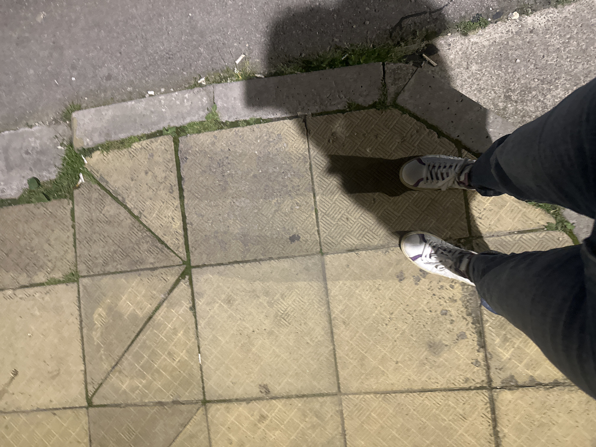 The image depicts a first-person perspective of a person standing on a paved area, possibly a sidewalk, next to a street. The paving consists of large, square, textured tiles in a yellowish-brown color, with visible grout lines. Some of these tiles are bisected by diagonal lines, creating triangular sections. Grass and weeds are growing in the gaps between the paving and the concrete curb, which separates the paved area from a gray asphalt street. The lighting suggests it is nighttime or a dimly lit environment, casting a noticeable shadow of the person's legs and feet onto the tiles. The person is wearing dark-colored pants and white sneakers with purple accents. Several discarded cigarette butts are visible on the curb and the edge of the street. The scene is consistent with an urban environment in Shumen, Bulgaria.