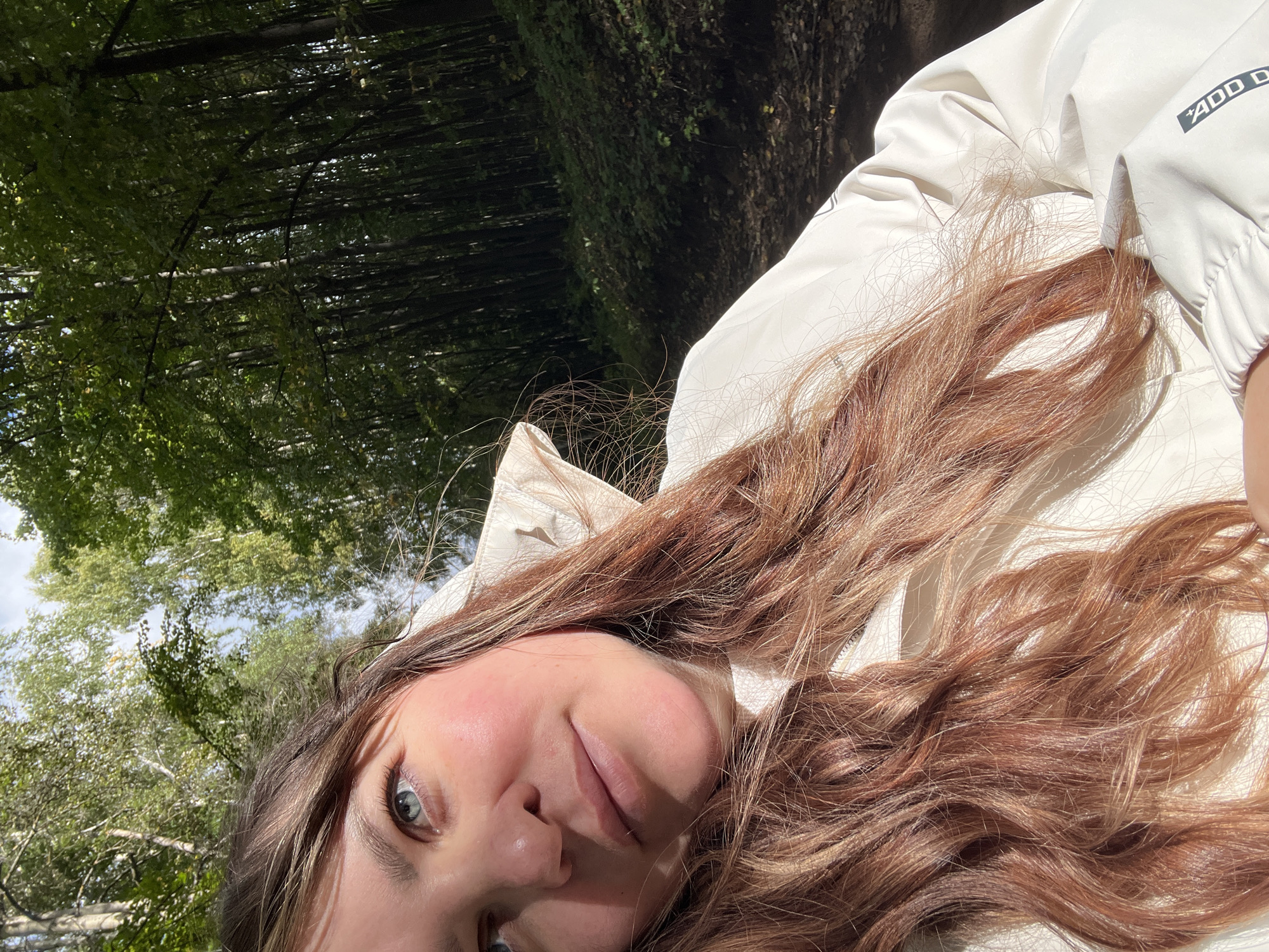 A young woman with shoulder-length, wavy auburn hair is captured in a close-up, selfie-style shot outdoors. She is wearing a light-colored hooded jacket with white lining visible around the neck and shoulders. The woman has light skin with a rosy blush on her cheeks and is looking slightly upward and to her left. Her blue eyes are bright, and her lips are slightly parted.
The background suggests a wooded area, with tall, slender trees and dense foliage. Sunlight filters through the leaves, creating dappled light and shadows on the ground, which appears to be a dirt path or trail. The overall atmosphere is natural and serene. There is no indication of specific landmarks or infrastructure that would pinpoint the location within Noviy, Russia, beyond it being a natural outdoor setting likely on the outskirts or within a park of the city. The image does not contain any objects or activities that would suggest an investigation or forensic relevance.