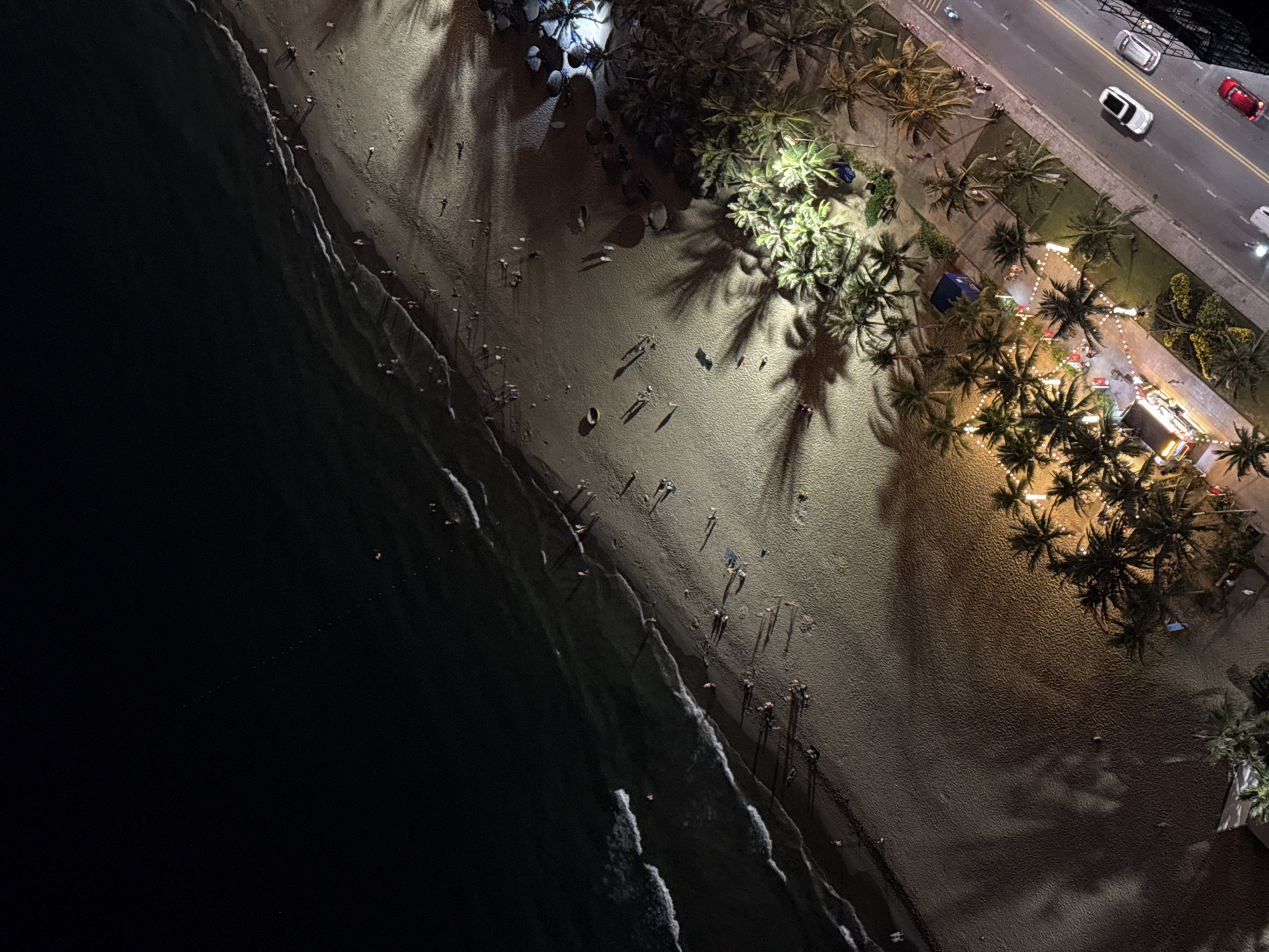 This is an aerial, nighttime photograph of a beach in Da Nang, Vietnam. The image captures a wide expanse of dark ocean meeting a sandy beach dotted with numerous people. Waves gently break along the shore. The beach is illuminated by artificial lights, casting long shadows from palm trees. Along the edge of the beach, there is a road with cars and pedestrians. Several food stalls or small businesses are visible, decorated with string lights. Palm trees line the area between the beach and the road.