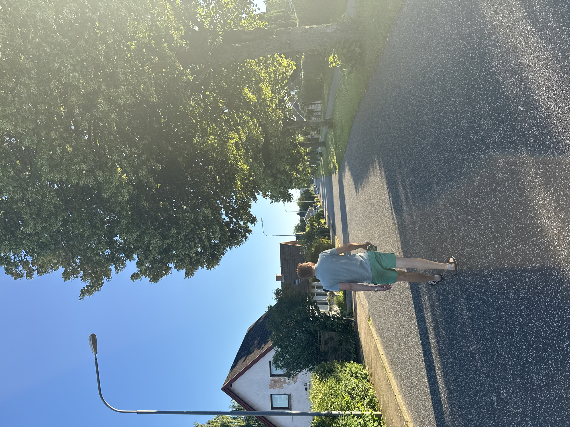 A man wearing a light blue t-shirt and green shorts is walking away from the camera down a paved residential street lined with trees and houses. He is wearing sandals and appears to be holding a clear bottle in his right hand. The street is asphalt with some white aggregate visible. The sun is shining brightly, casting long shadows across the road. The sky is clear and blue. On the left side of the street, there are houses with pitched roofs, hedges, and trees. On the right side of the street, there are more houses and a sidewalk, also bordered by trees and vegetation. The scene appears to be a suburban area, likely in Vejle, Denmark, given the architectural style of the houses and the presence of streetlights.