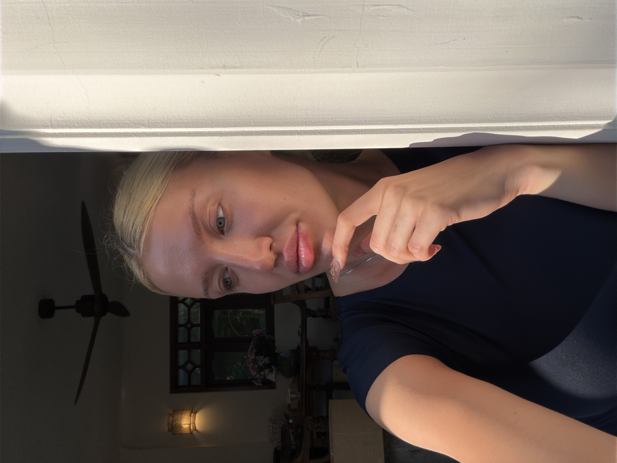 A young woman with blonde hair and blue eyes is positioned in the foreground, leaning against a textured white column. She is looking to her left, with her right hand resting on her chin. She is wearing a dark blue t-shirt. The background is dimly lit, showing a ceiling fan, a decorative lamp, and a table with a vase of flowers. The scene appears to be indoors, possibly on a veranda or patio in North Kuta, Indonesia, given the presence of a ceiling fan, likely in a residential or hospitality setting. The lighting suggests natural daylight entering from the left side of the frame.
