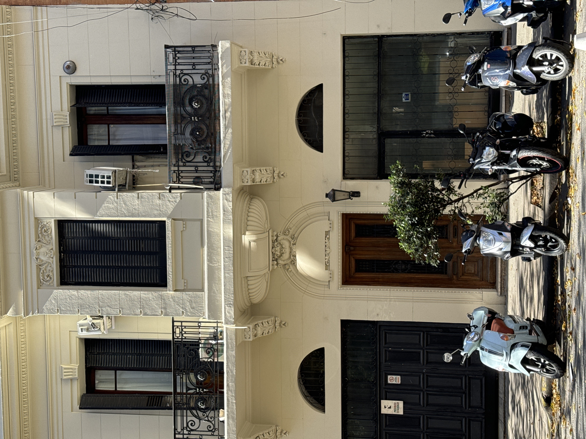 The image shows the facade of a building in Buenos Aires, Argentina. The building has three stories, each with a window and balcony. The windows are all shuttered. The facade is light-colored with ornate architectural details. There are several scooters parked on the sidewalk in front of the building. The street is paved with asphalt and lined with trees. The photo is taken during daylight hours.