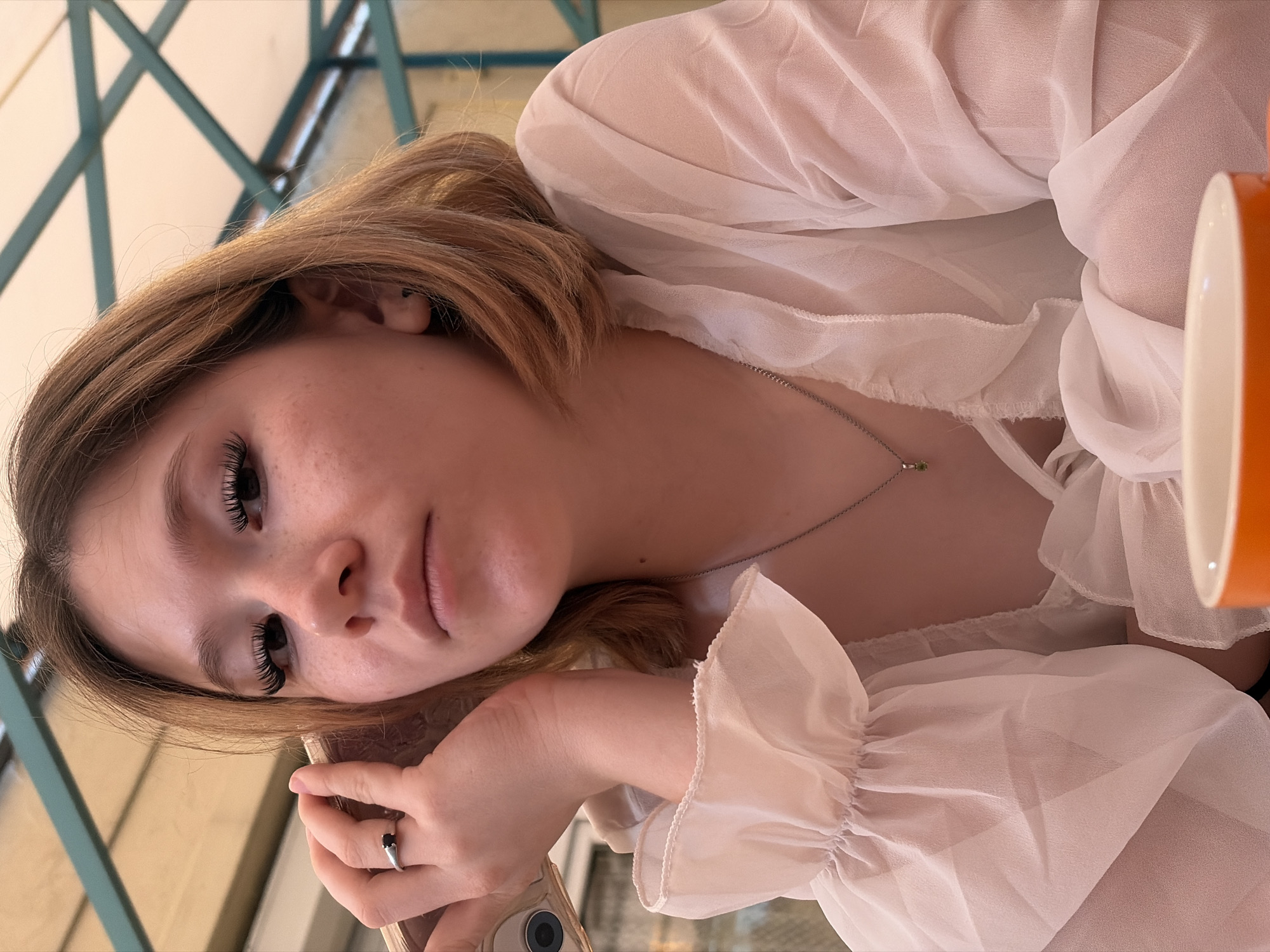 The image shows a close-up, portrait-oriented shot of a young woman. She has light brown hair that is styled loosely around her face. Her eyes are open and she is looking towards the viewer with a neutral expression. She appears to be resting her head on her arm, and her hand is visible, wearing a ring on one finger. She is wearing a sheer, white blouse with ruffled sleeves. A delicate silver necklace with a small green pendant is visible around her neck. To the right of the frame, a portion of an orange object, possibly a mug or cup, is visible. The background is blurred, but appears to be an outdoor setting with some metallic structures, possibly a patio or balcony. The lighting suggests it could be daytime.