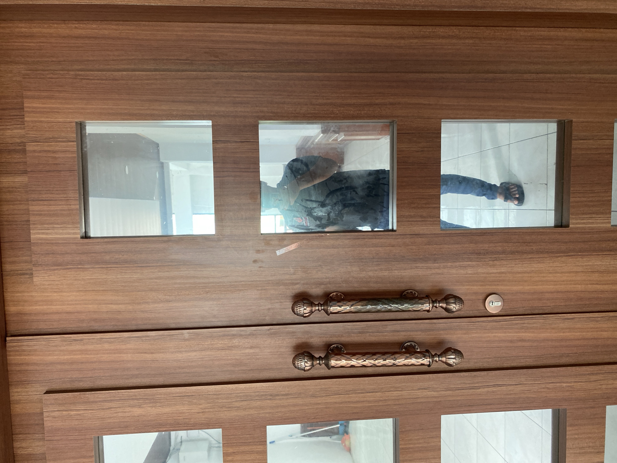 The image shows a close-up view of a wooden double door with glass panes. The door has two large, ornate copper handles and a keyhole. The wood grain of the doors is prominent, running vertically. Reflected in the glass panes are images of a person. The person is wearing a dark t-shirt and blue jeans. The environment reflected in the glass suggests an indoor setting with tiled floors and walls. The overall impression is of a modern, well-maintained entrance. The location is identified as Banda Aceh, Indonesia.