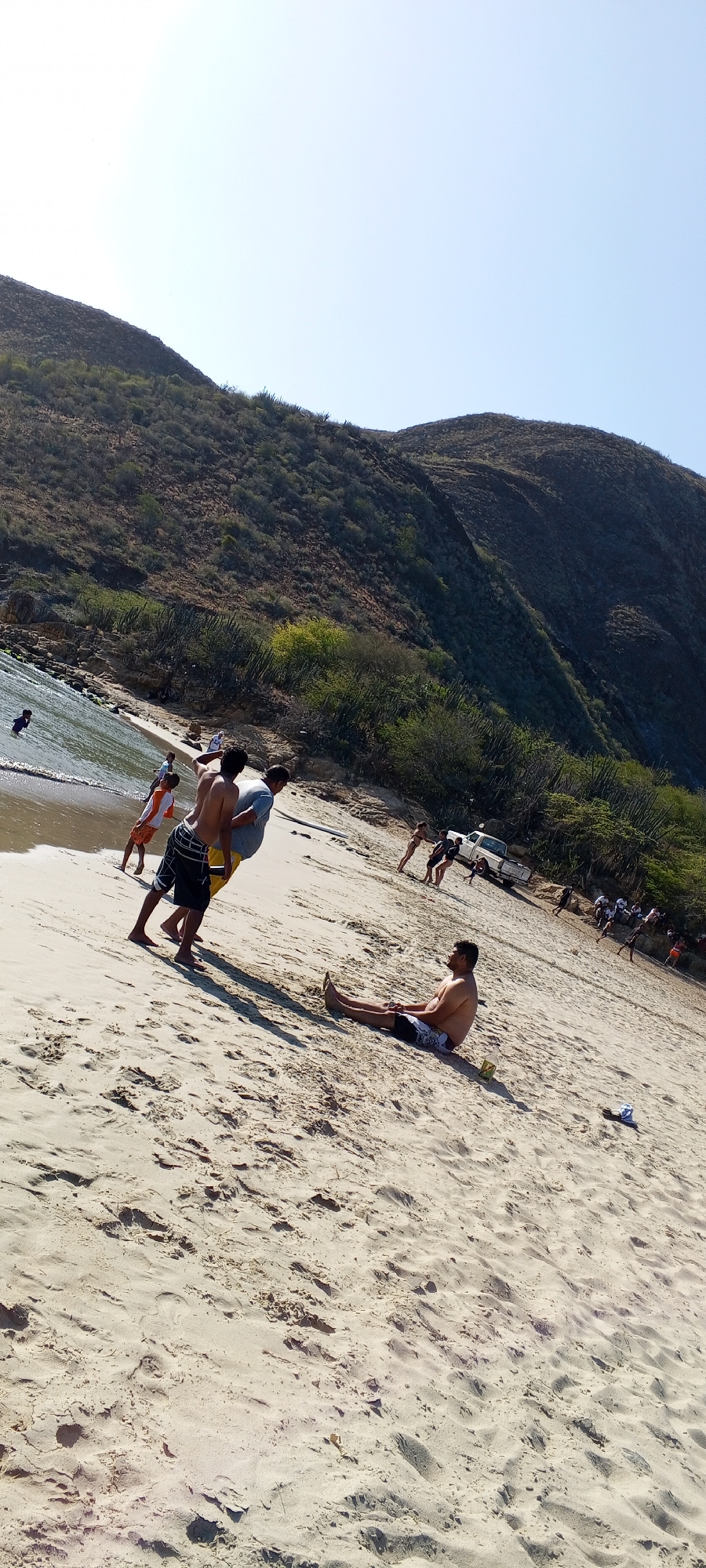 The image shows a beach scene on a sunny day. In the foreground, a man sits on the sand. Two men stand nearby, one holding up the other. Further in the background, several people are playing near the water's edge, and a white pickup truck is parked close to the vegetation line. Behind the beach, a large, hilly landscape rises into the sky. The image was likely taken in an outdoor location given the sand and water.