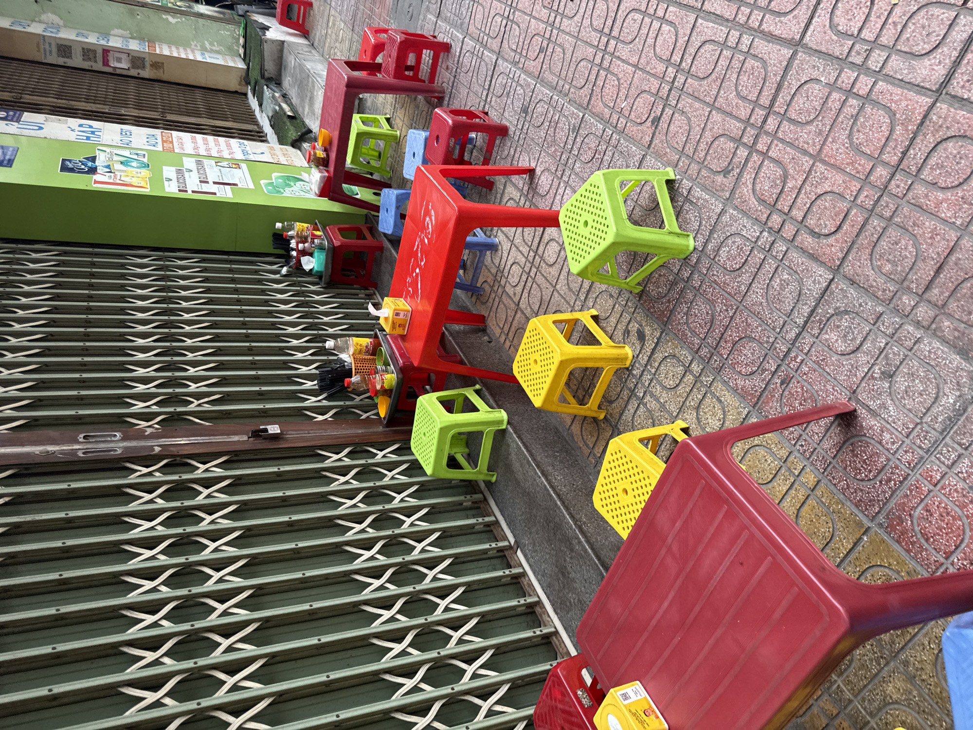 This is a photograph of an outdoor dining area located on a sidewalk in Nha Trang, Vietnam. The scene features a collection of brightly colored plastic tables and stools, typical of street food vendors in the region. A green metal roll-up gate, commonly used for storefront security, dominates the background. The pavement is tiled with a patterned design in shades of red and beige. Scattered on the tables are various condiments, bottles, and paper napkins, suggesting the space is ready for customers. There are no people visible in the frame.