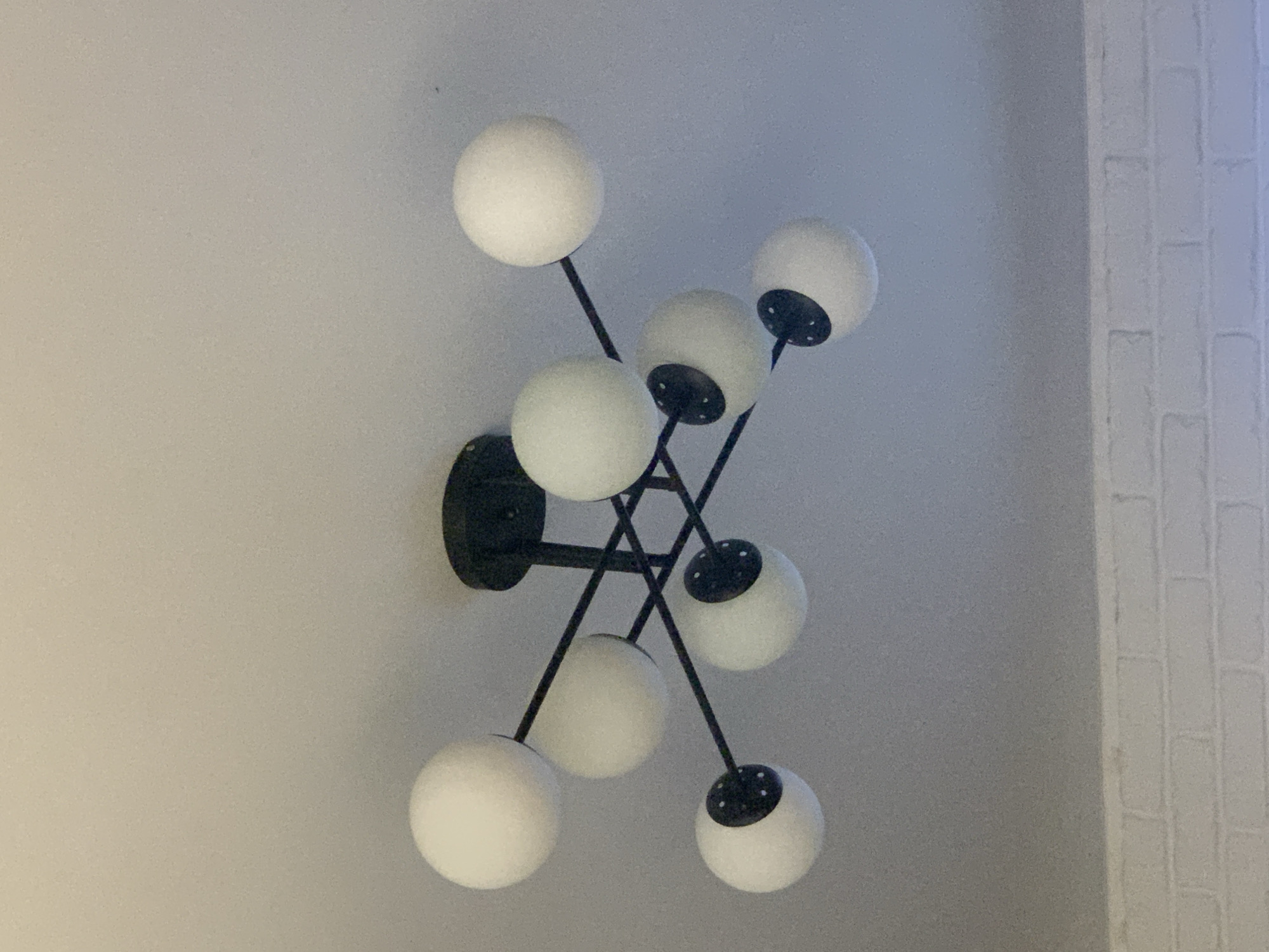 A black, multi-arm chandelier with nine white globe-shaped lights is mounted on a white ceiling. The chandelier has a central black base from which thin black rods extend, supporting the lights at various angles. Some of the lights have small black circular accents on the bottom. The ceiling appears to be a flat, painted surface, possibly textured. Below the ceiling, a portion of a white brick wall is visible. There are no people or animals in the image. The image was taken indoors.