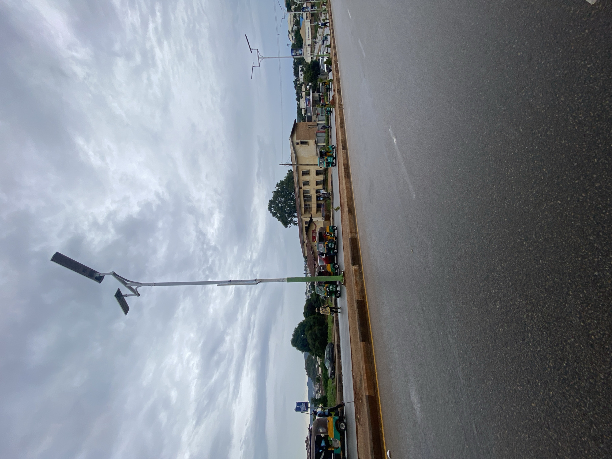 An overcast day in Jos, Nigeria, is depicted in this photograph, which features a wet asphalt road. The road is lined with a concrete sidewalk and a planter area. Several three-wheeled vehicles, commonly known as Keke Napeps, are visible, as are a few pedestrians. In the background, there are buildings, trees, and a hillside. A tall streetlamp with two solar panels stands prominently in the foreground.