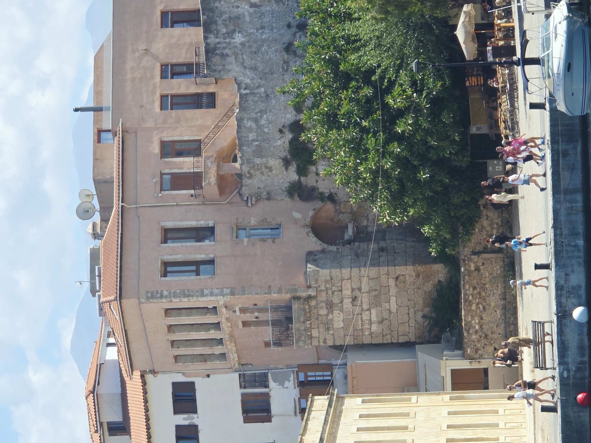 The image depicts a view of the Venetian Harbor in Chania, Greece. In the foreground, a waterfront promenade is visible with several people walking. A light-colored building with arched windows stands on the left, and a stone wall with an arched opening is on the right. Behind these structures, a multi-story building with a light pink facade and terracotta roof tiles is prominent. The sky is blue with scattered clouds, and distant mountains are visible. A boat is docked in the water in the lower right corner. The scene suggests a sunny day in a historic coastal city.