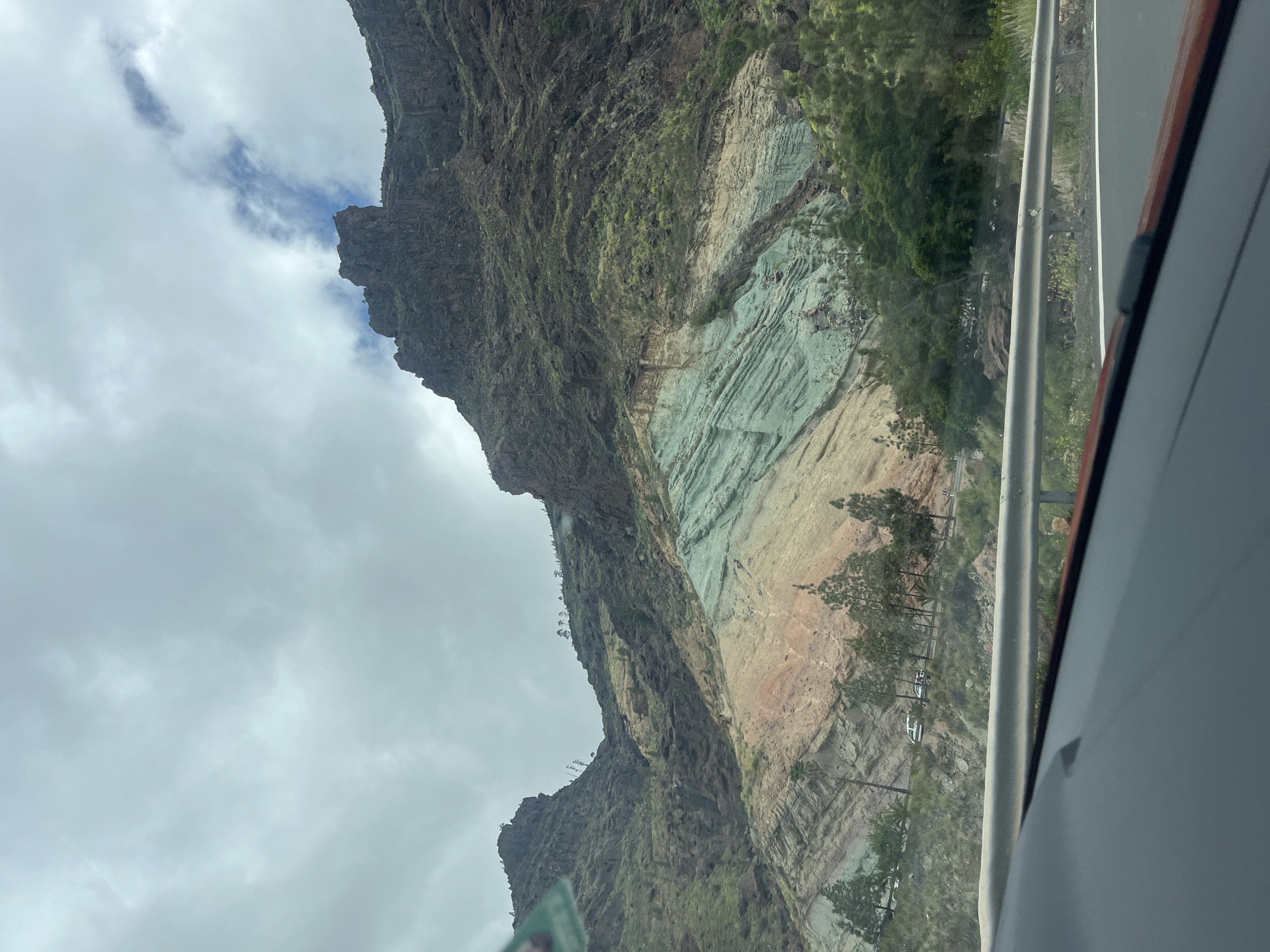 The image, taken from inside a vehicle, shows a mountainside in Mogán, Spain. The mountain exhibits distinct geological layers, with bands of dark brown, light brown, and a striking turquoise green. Vegetation clings to the slopes. A road runs alongside the mountain, partially obscured by the vehicle's interior. The sky is overcast with grey clouds.