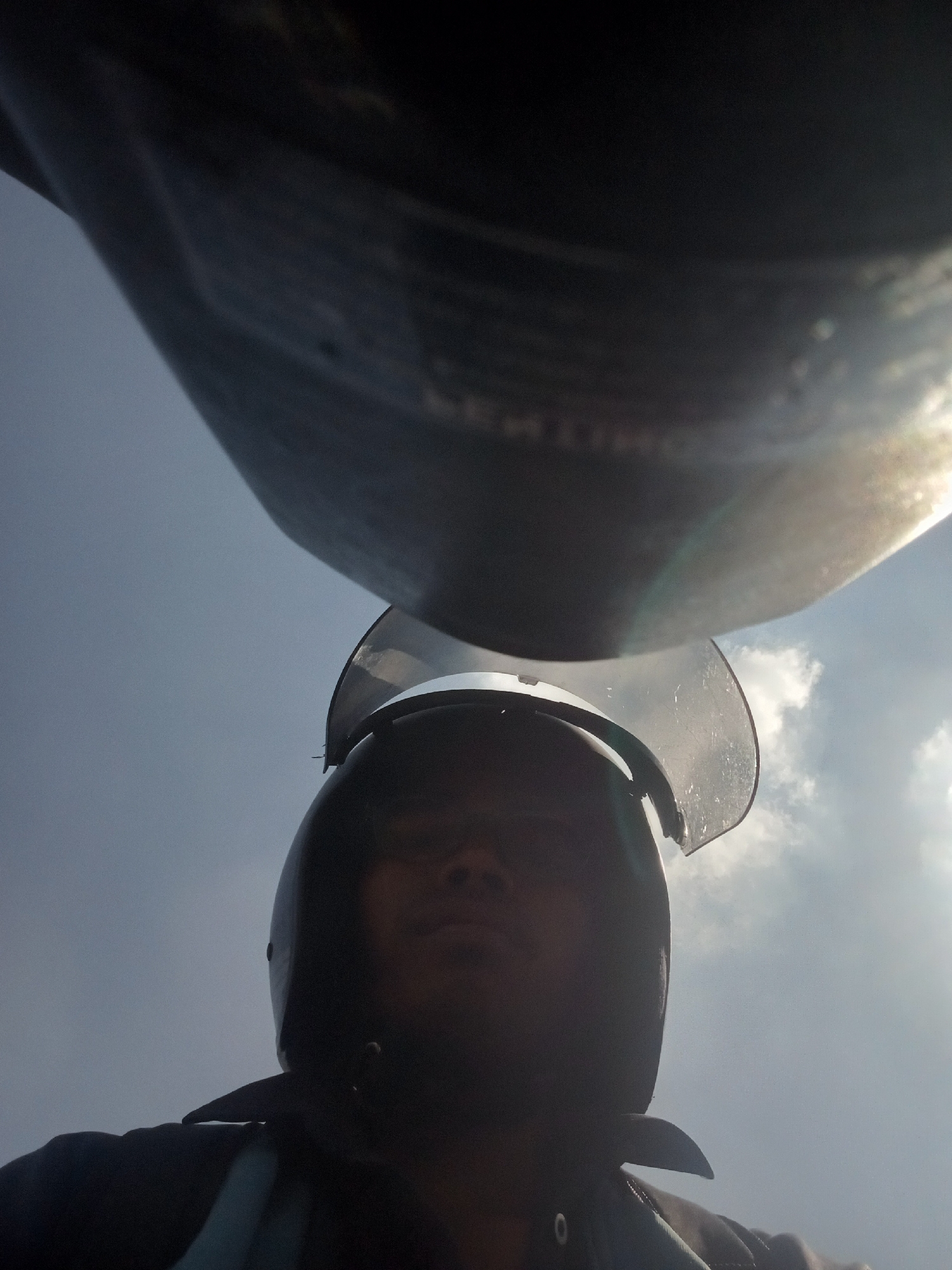 A low-angle, close-up, point-of-view shot shows a person wearing a dark helmet with a clear face shield, looking up towards the sky. A large, dark, curved object, possibly part of a vehicle or structure, looms overhead, partially obscuring the bright, cloudy sky. The person appears to be outdoors in a bright, sunny environment. The context suggests the image might have been taken in Surabaya, Indonesia, given the user's prompt.