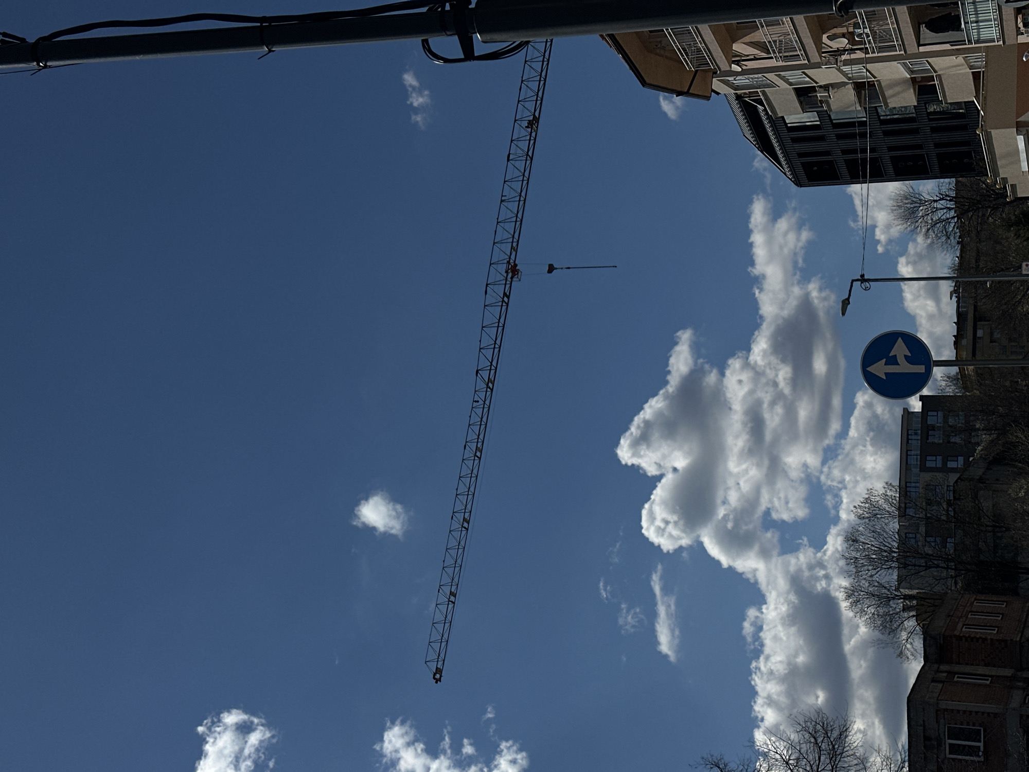 The image depicts a low-angle view of the sky in an urban environment, likely Sofia, Bulgaria. A tall construction crane is visible against a clear blue sky dotted with white clouds. Adjacent to the crane, there is a multi-story building with balconies and windows. In the lower right corner, a blue traffic sign with white arrows indicating a fork in the road is mounted on a pole. Bare trees and other buildings, likely residential, are also partially visible. Power lines run across the top portion of the image.