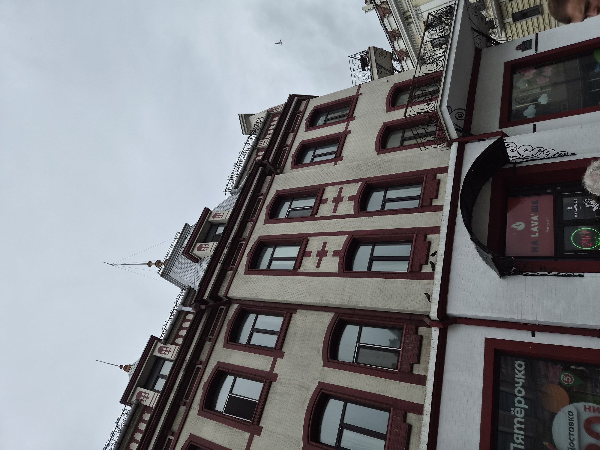 A low-angle, eye-level medium shot captures the facade of a multi-story building in Vladivostok, Russia, under an overcast sky. The building features cream-colored brickwork with dark red trim accentuating the windows, architectural details, and corners. The building's roofline is complex, with gabled sections and decorative finials. Several pigeons are perched on the building's ledges. On the ground floor, signage indicates commercial establishments, with one sign clearly reading "Пятёрочка" and another displaying "HA LAVA'ШЕ ROASTER CAFE." A portion of a person's head is visible in the lower right corner of the frame. The architectural style suggests an older, possibly historical, building in an urban setting.