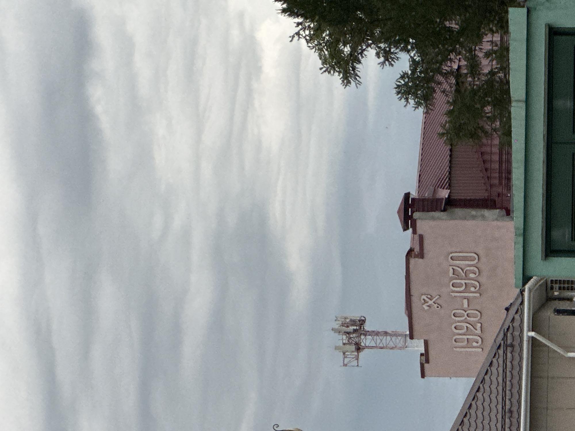 The image shows the facade of a building with a large sign that reads "1928-1930". Above the numbers is a symbol resembling a hammer and sickle, often associated with communism. The building appears to be made of concrete and painted a pale pink color. To the left of the main building, a communication tower with antennas is visible, and to the right, a dense green tree partially obstructs the view of another building with a red metal roof. In the foreground, a portion of a brown-tiled roof is visible, suggesting this is a low-rise area. The sky is overcast with grey clouds. This building is located on Volodarskogo Street in Omsk, Russia.