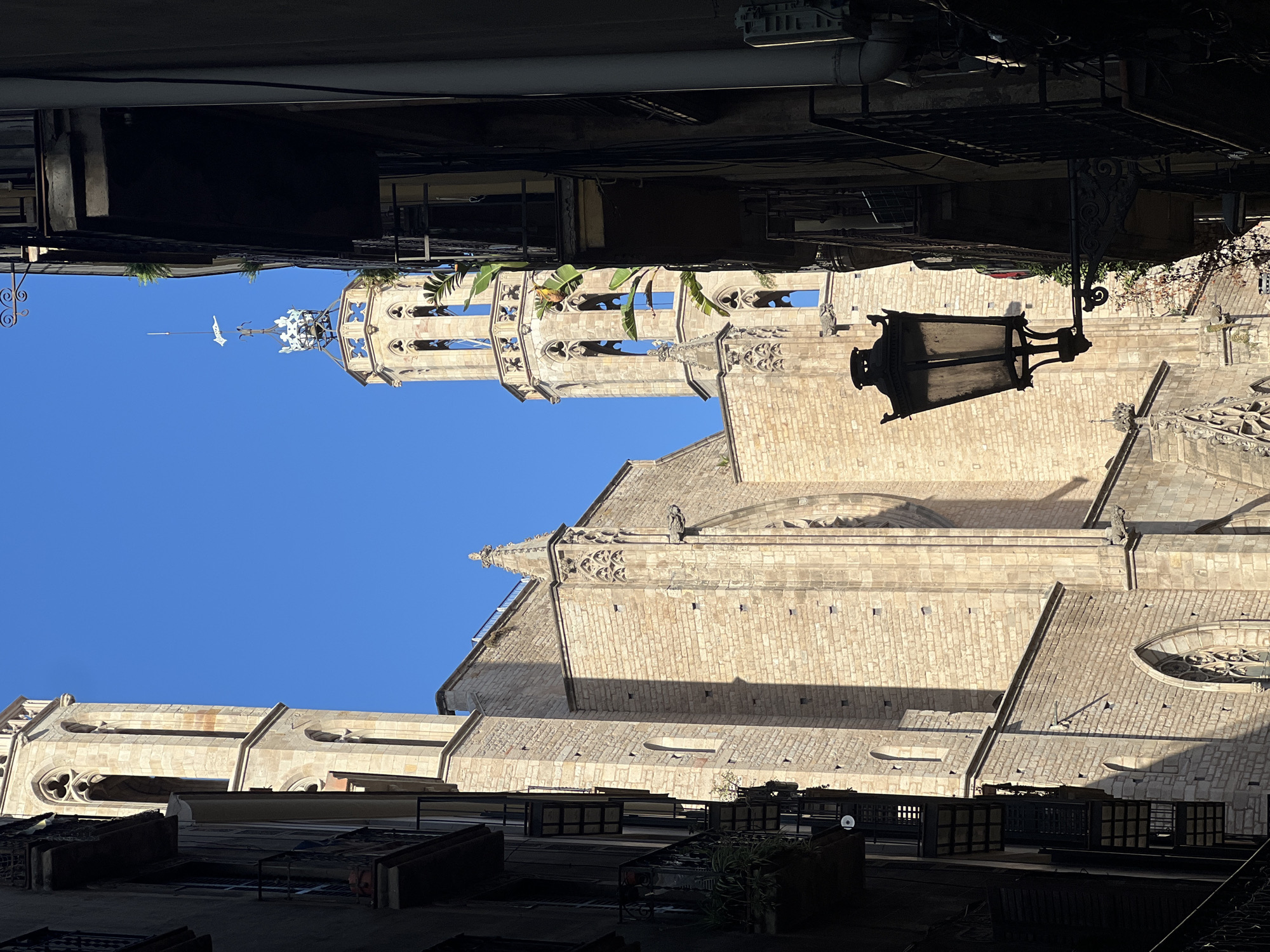 This image captures a narrow urban street in the Gothic Quarter of Barcelona, Spain, providing a view of the **Barcelona Cathedral (Catedral de la Santa Creu i Santa Eulàlia)**.
The dominant subject is the cathedral, constructed from light-colored stone blocks, with its intricate Gothic architecture. Two bell towers are visible against a clear, deep blue sky. The taller, more prominent tower on the right features multiple tiers of arched openings, ornate tracery, a distinct crown-like architectural element, and a finial topped with a weathervane. A second, partially visible tower stands to the left. The cathedral's side wall, extending from left to right, displays buttresses, arched windows, and a gargoyle protruding near a decorated pinnacle. Strong sunlight illuminates portions of the cathedral's facade, creating deep shadows along its left side and within architectural recesses.
Flanking the narrow street on both the left and right are dark, multi-story buildings, casting significant foreground shadows. These structures feature balconies with wrought iron railings, some adorned with potted plants. A large, dark, ornate streetlamp with an attached scrolled bracket extends from the right-hand building into the street space. No individuals are present in the scene.