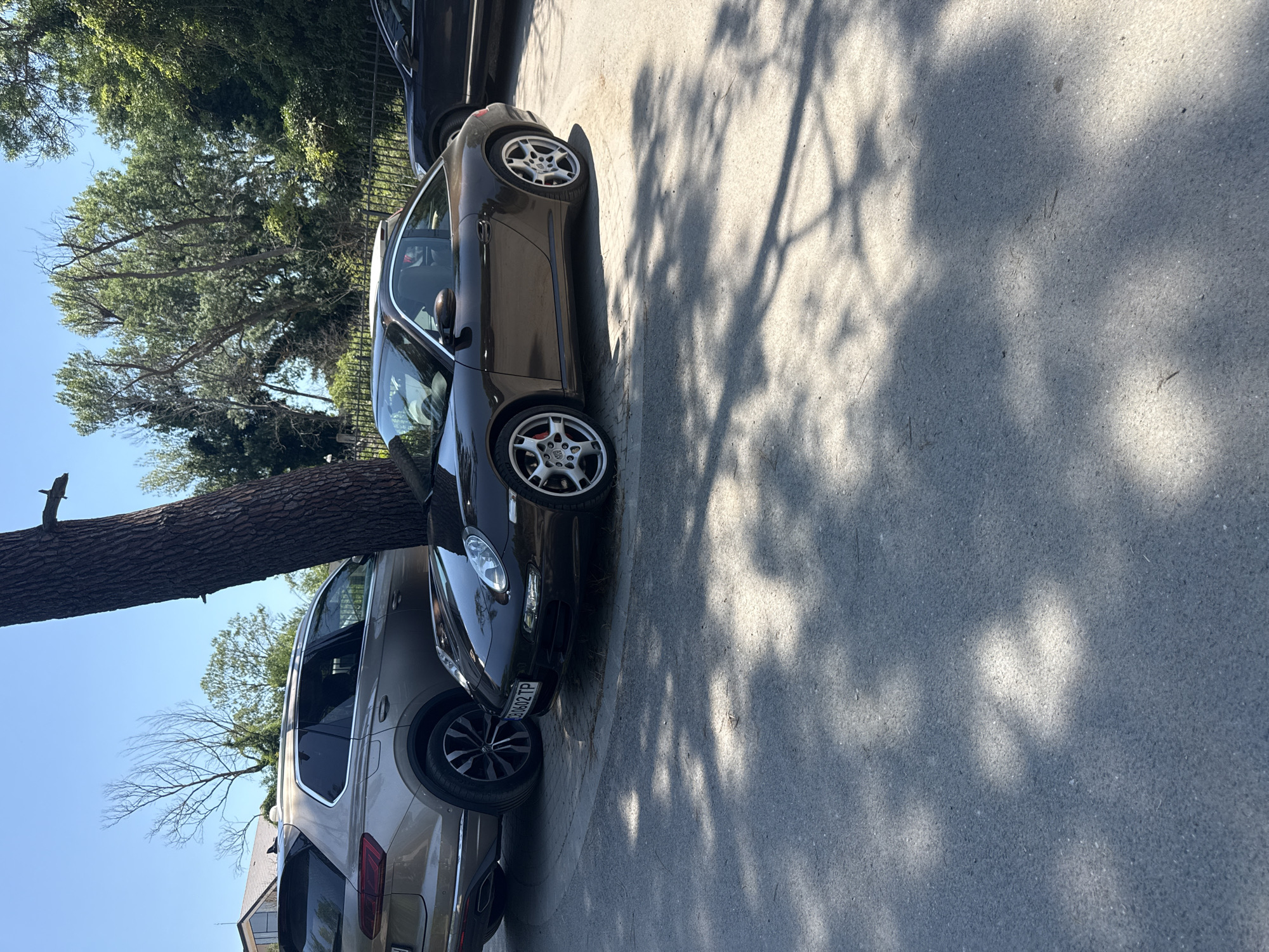 A dark brown Porsche 911 coupe is parked on an asphalt surface next to a tan SUV. The Porsche's front license plate reads "CB 0602 TP". In the background, there are trees, a black fence, and a building. The scene appears to be outdoors on a sunny day with dappled sunlight and shadows on the ground. The location is identified as Dyuni, Bulgaria.