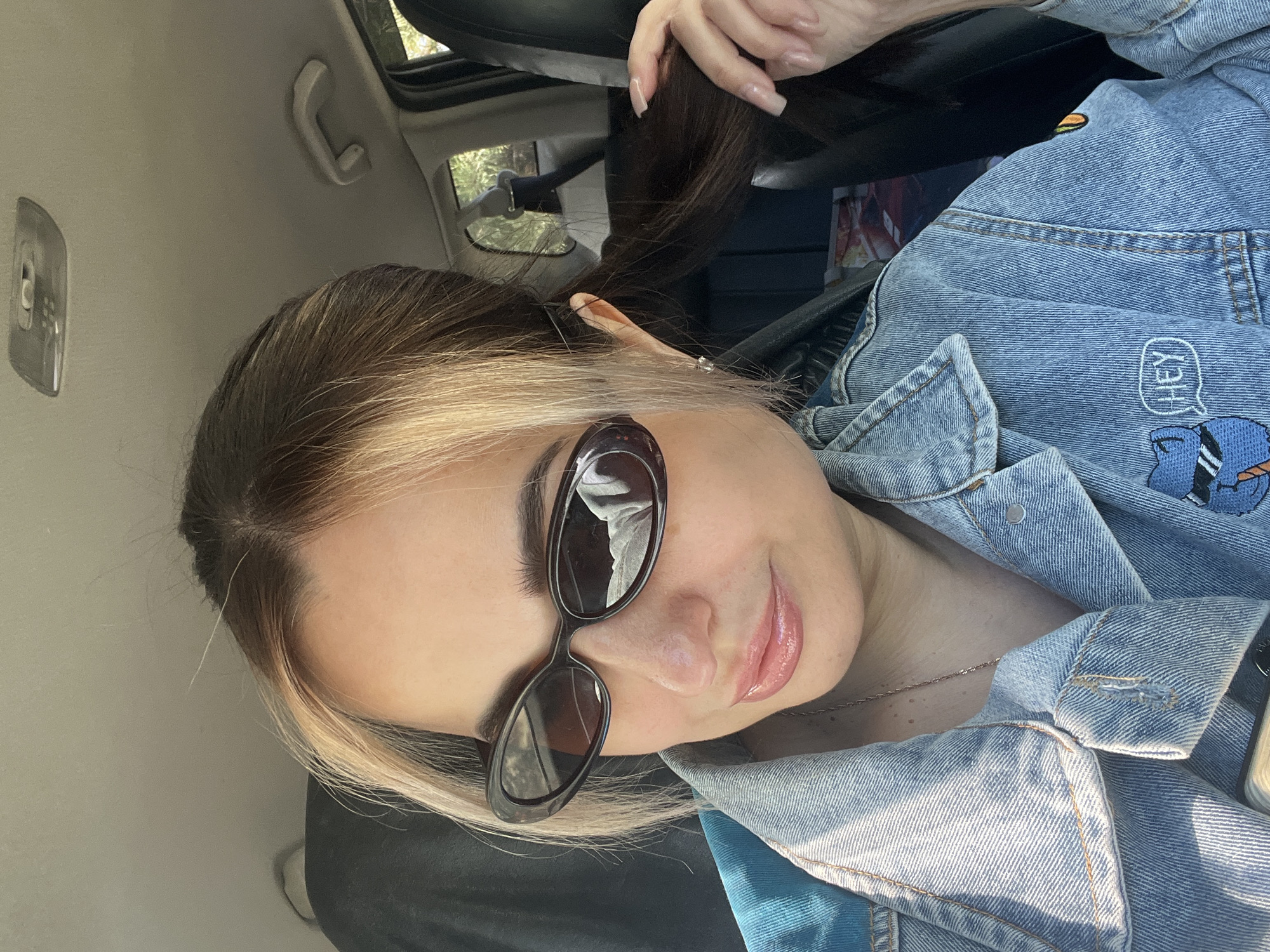 A woman is seated inside a car. She is wearing sunglasses and a denim jacket with a patch on the left side. She has brown hair with blonde highlights and is holding her ponytail with her right hand. The car's interior is visible, including the sunroof and a light fixture. The location is Unknown, Unknown.
